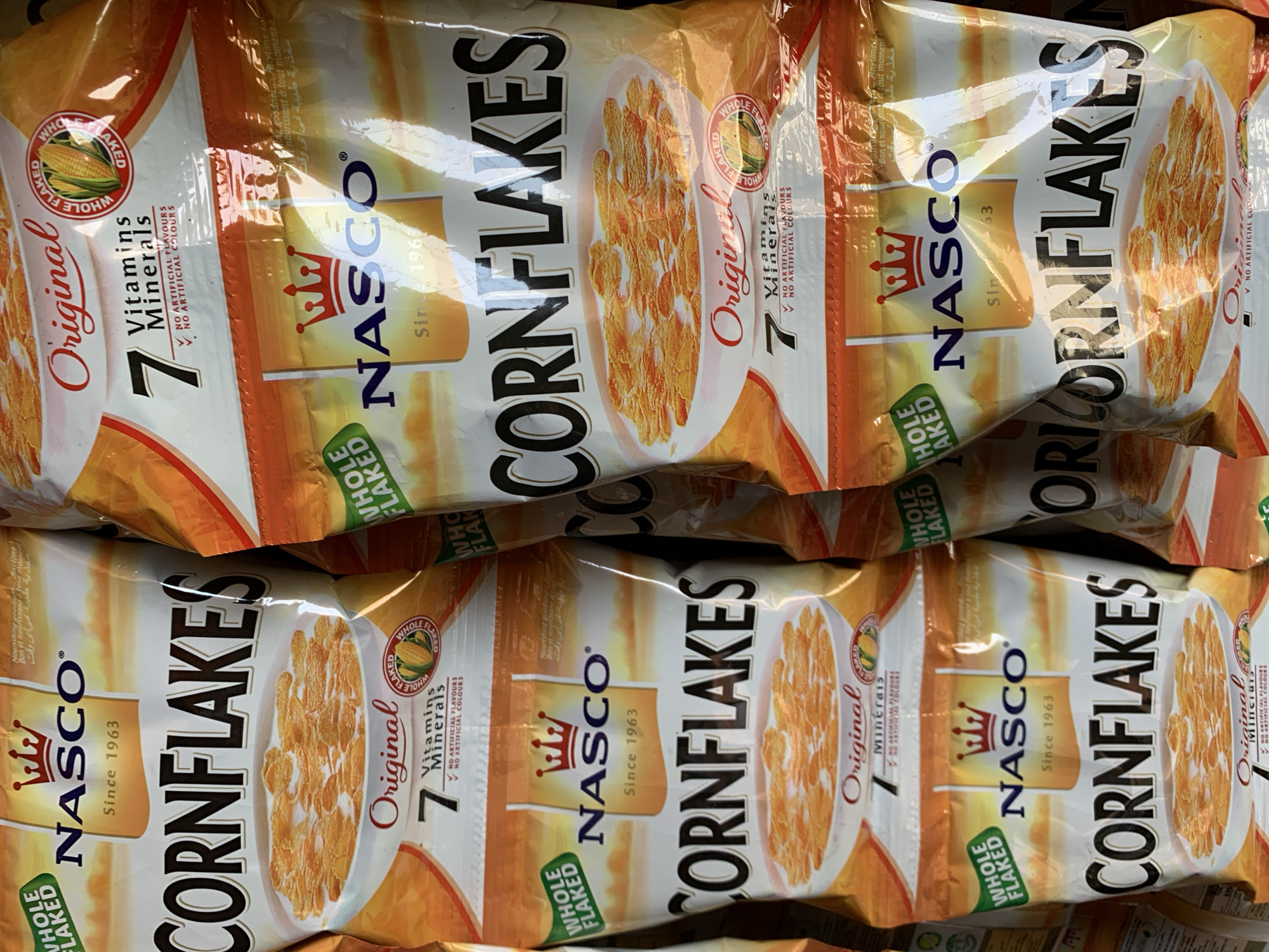 The image shows a close-up view of multiple packages of "NASCO Cornflakes." The packages are predominantly white and orange, with bold black lettering for "CORNFLAKES." The packaging also includes the "NASCO" logo with a crown, the tagline "Since 1963," a bowl of cornflakes, a corn stalk graphic, and text indicating "7 Vitamins Minerals" and "No Artificial Flavours/Colours." The packages appear to be stacked in a retail environment, possibly in Jos, Nigeria, given the brand's history and prevalence in the region.