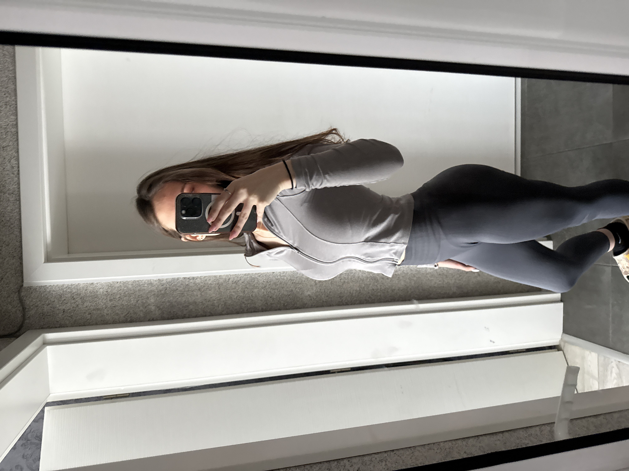 A woman is taking a mirror selfie in what appears to be a home. She is wearing grey leggings and a grey zip-up jacket. She is holding a smartphone in her left hand, partially obscuring her face. Her brown hair is long and loose. The background includes a white door frame, light grey carpet, and what looks like a wall with grey tiles. The location in the city of Sofia, Bulgaria, cannot be determined from the image.