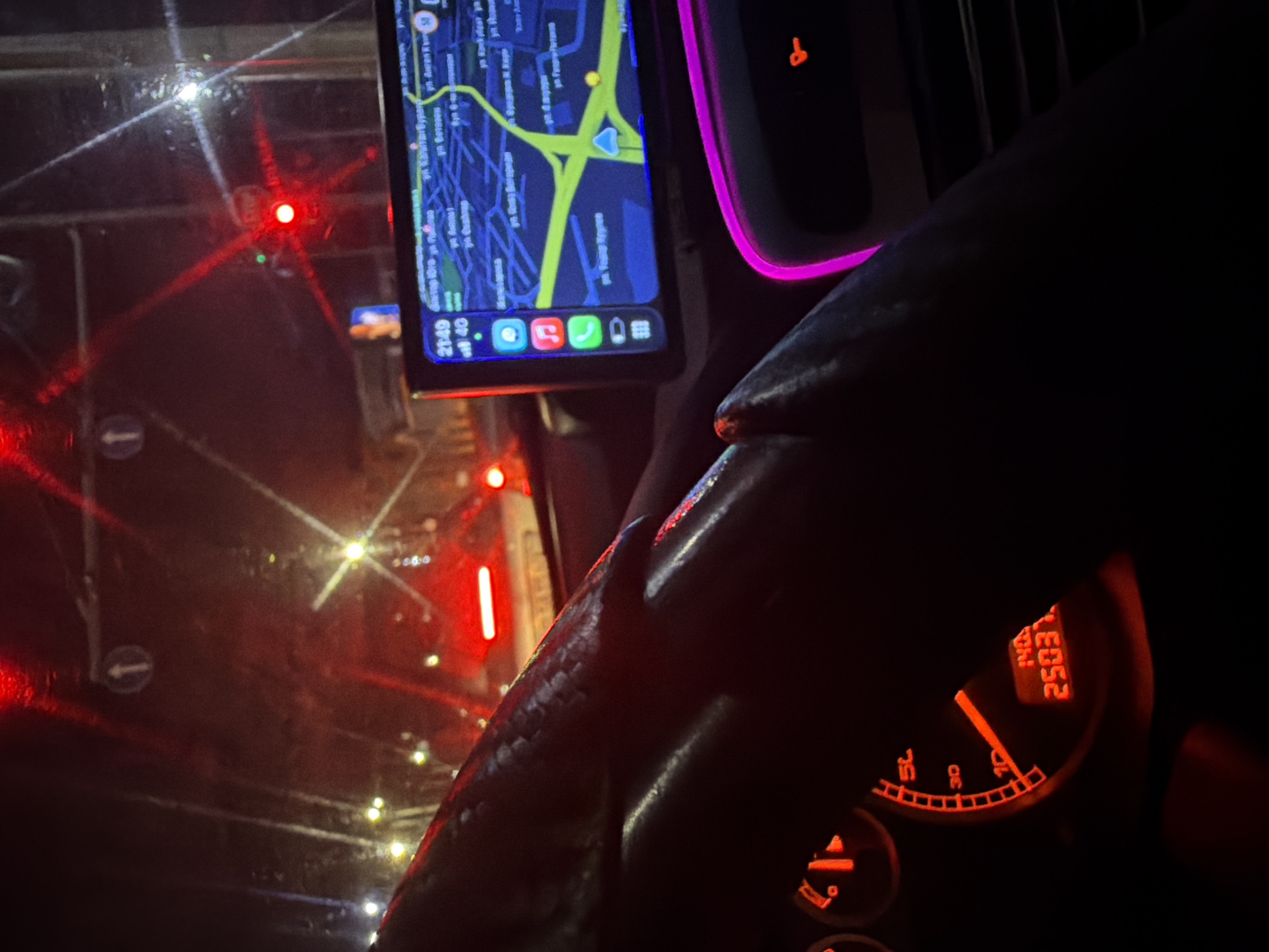 The image shows a first-person perspective of a driver inside a car at night, likely in Plovdiv, Bulgaria. The driver's hand is on the steering wheel, which is partially visible in the foreground. The dashboard lights are illuminated with a red glow, including a speedometer and an odometer displaying "25031". A GPS navigation screen is mounted on the dashboard, showing a map with street names in Cyrillic. Outside the car, city lights and traffic lights are visible through the windshield, with a red traffic light indicating the car is stopped. The windshield appears wet, suggesting rain or moisture. The overall scene depicts a night-time drive through a city, possibly in traffic.
