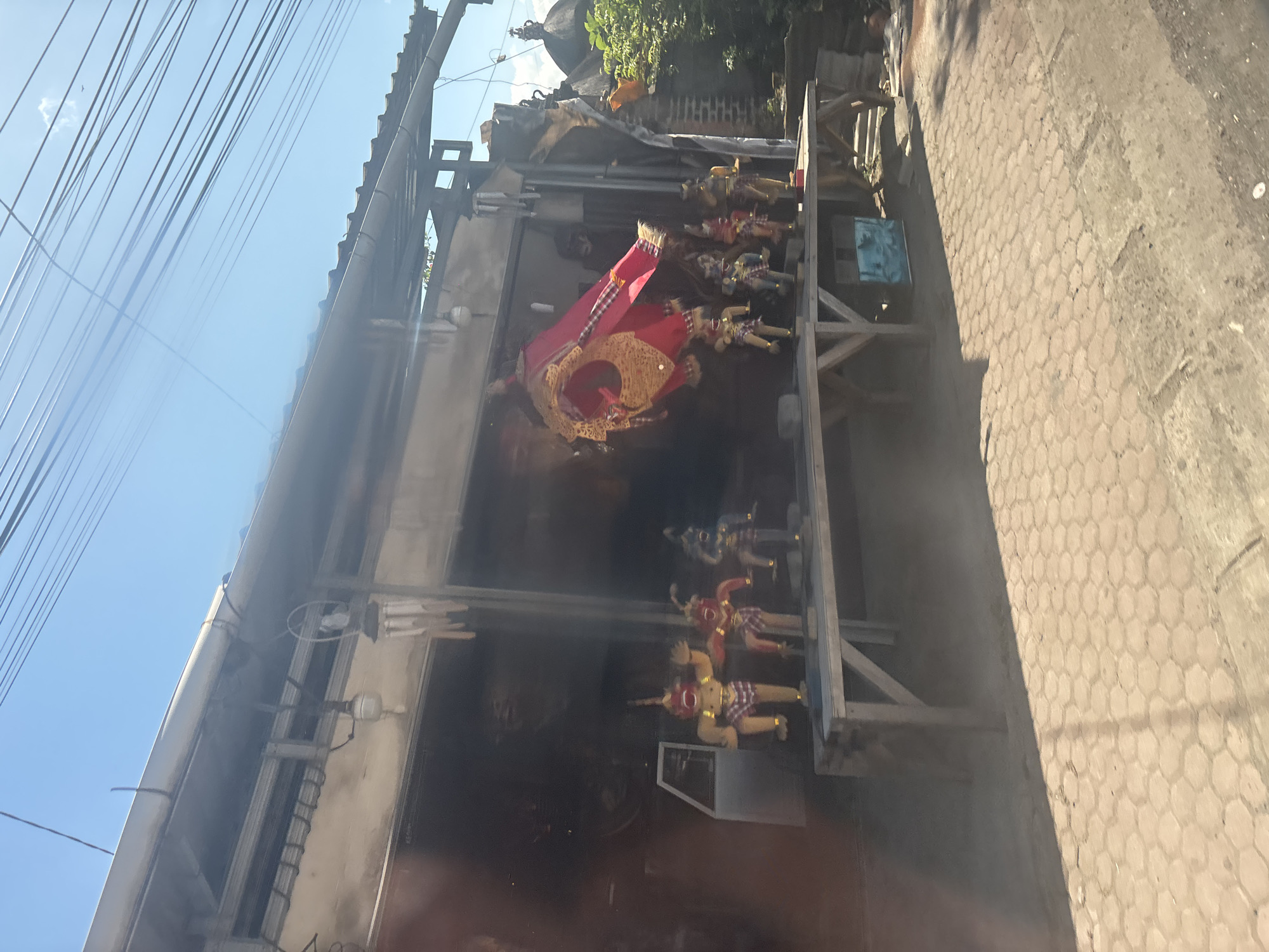 The image depicts a street-level view of a shop or stall in Sukawati, Indonesia, on a sunny day. The focal point of the image is a display of traditional Balinese puppets, likely for sale. Several figures, characterized by their distinctive costumes and elaborate headdresses, are arranged on a wooden table in front of the shop. One large, red and gold puppet, possibly a representation of a mythical creature or deity, is suspended above the smaller figures. The shop itself appears to be an open-air structure with a corrugated metal roof supported by beams. Behind the puppets and display table, glimpses of the shop's interior can be seen, including what looks like a white vehicle. The ground in front of the shop is paved with hexagonal interlocking stones. Overhead, a tangle of power lines crisscrosses the bright blue sky. The scene suggests a typical commercial area in Sukawati, known for its art markets and crafts.
