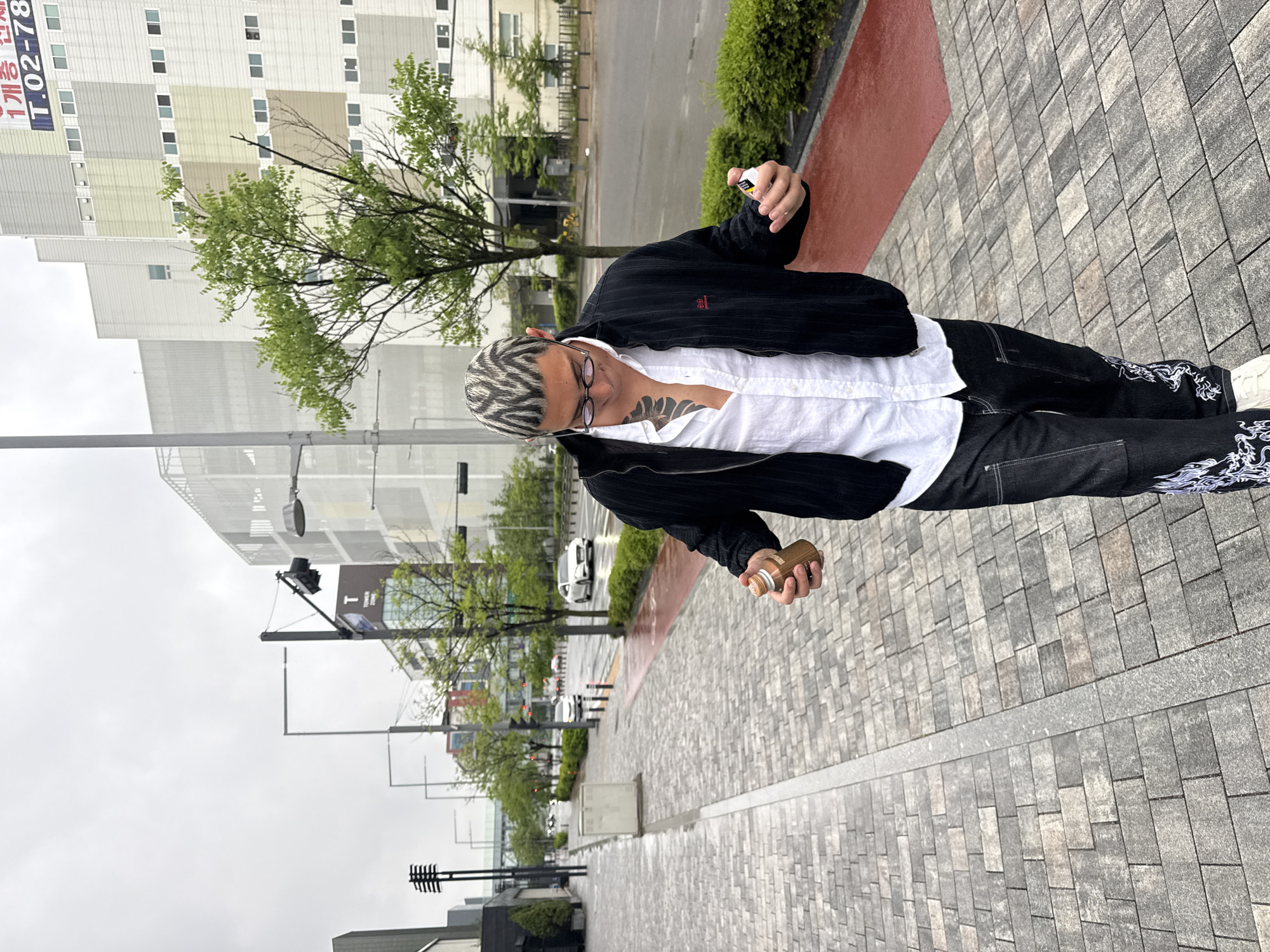A man is on a sidewalk in Gimpo-si, South Korea, in front of a building with trees. He is wearing a black jacket, a white shirt, and black pants with white designs. He has zebra-striped hair and is wearing glasses. He is holding a spray can in each hand.