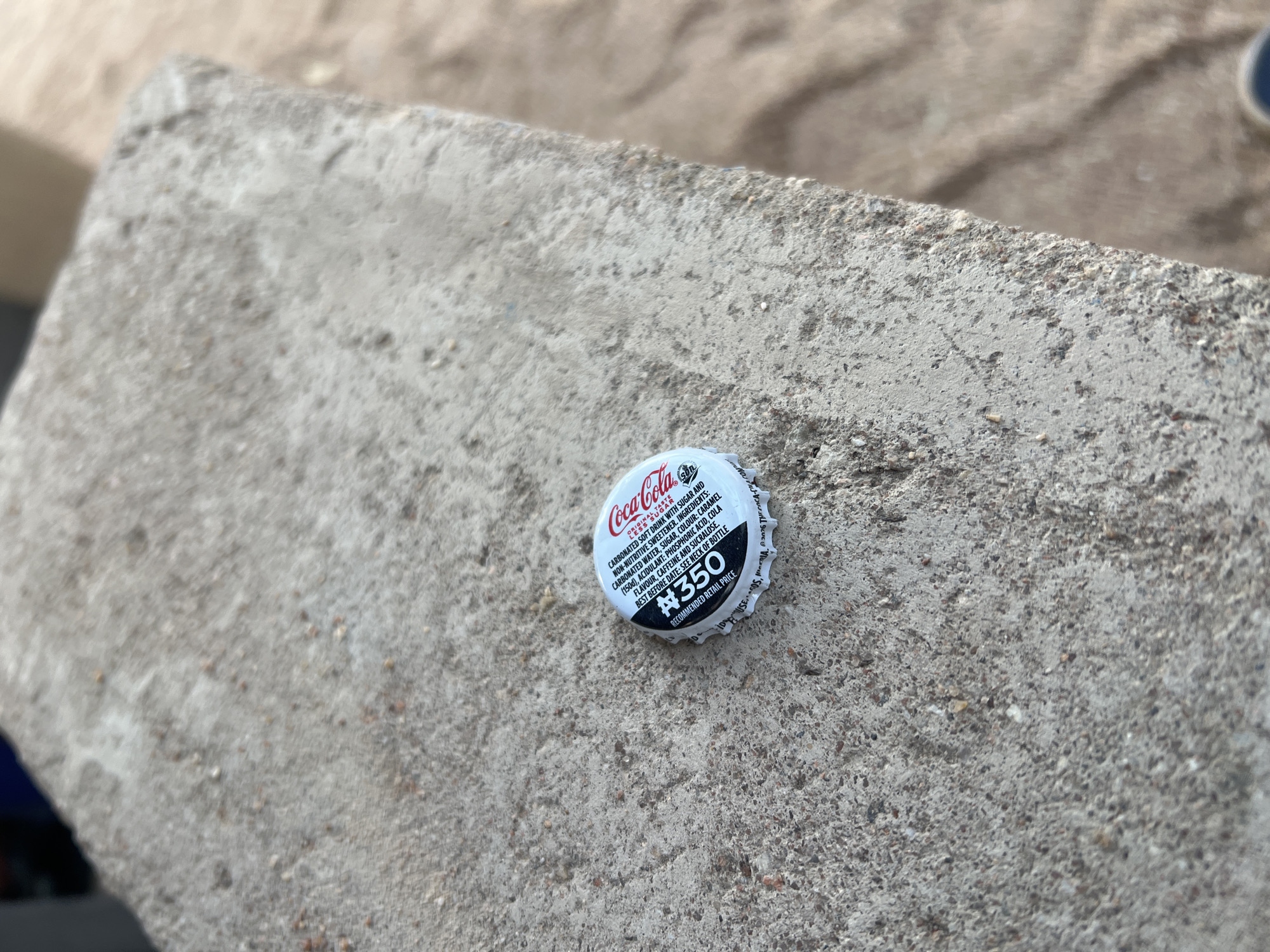 This media file is a close-up shot of a Coca-Cola bottle cap resting on a textured, grey concrete surface. The cap is white with red lettering and displays the iconic Coca-Cola logo. Visible text on the cap includes "Coca-Cola ORIGINAL TASTE", a list of ingredients, a "BEST BEFORE DATE", and the price "#350 RECOMMENDED RETAIL PRICE". The background is slightly blurred, showing more of the concrete surface and a hint of a beige, sandy-looking area. The lighting suggests it could be daytime, with a neutral to slightly overcast sky given the even illumination and lack of strong shadows. The image focuses on the discarded bottle cap, suggesting a scene of everyday life or perhaps a casual outdoor setting. No people are visible. The location context of Jalingo, Nigeria, implies the currency symbol "#" refers to the Nigerian Naira.