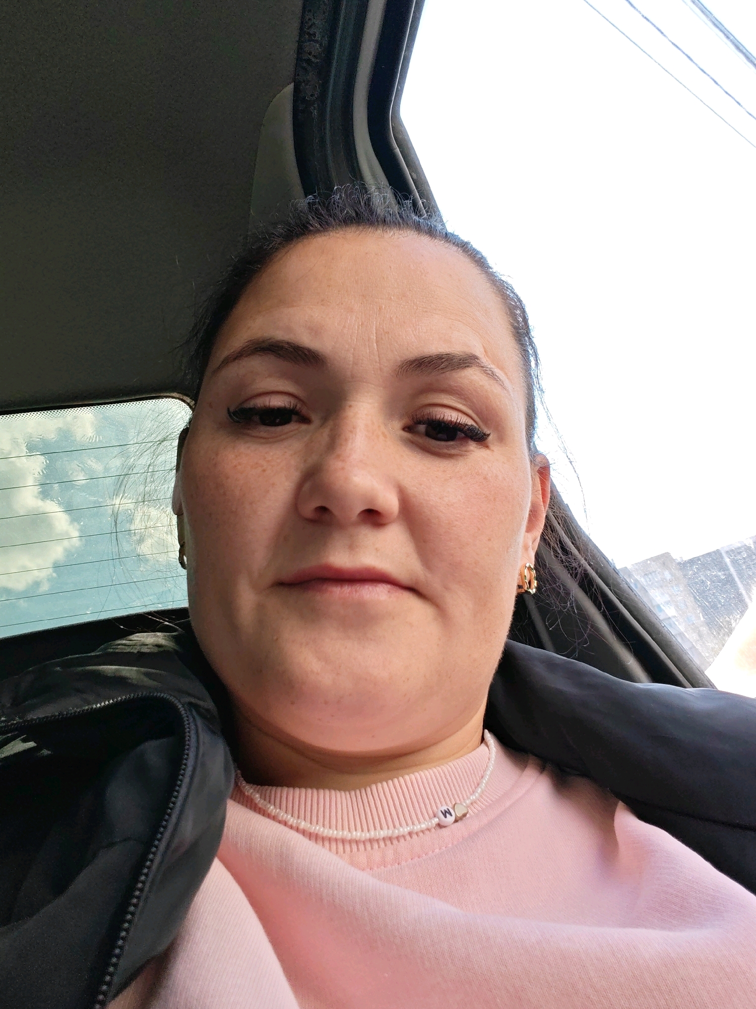 A woman is shown from the chest up, indoors, possibly in a vehicle. She has brown hair pulled back, brown eyes, and freckles on her face. She is wearing a black jacket over a pink sweatshirt and a pearl necklace with a small heart charm. The background appears to be a window with a cloudy sky visible. This image does not contain any information that would allow it to be located in Perm, Russia.