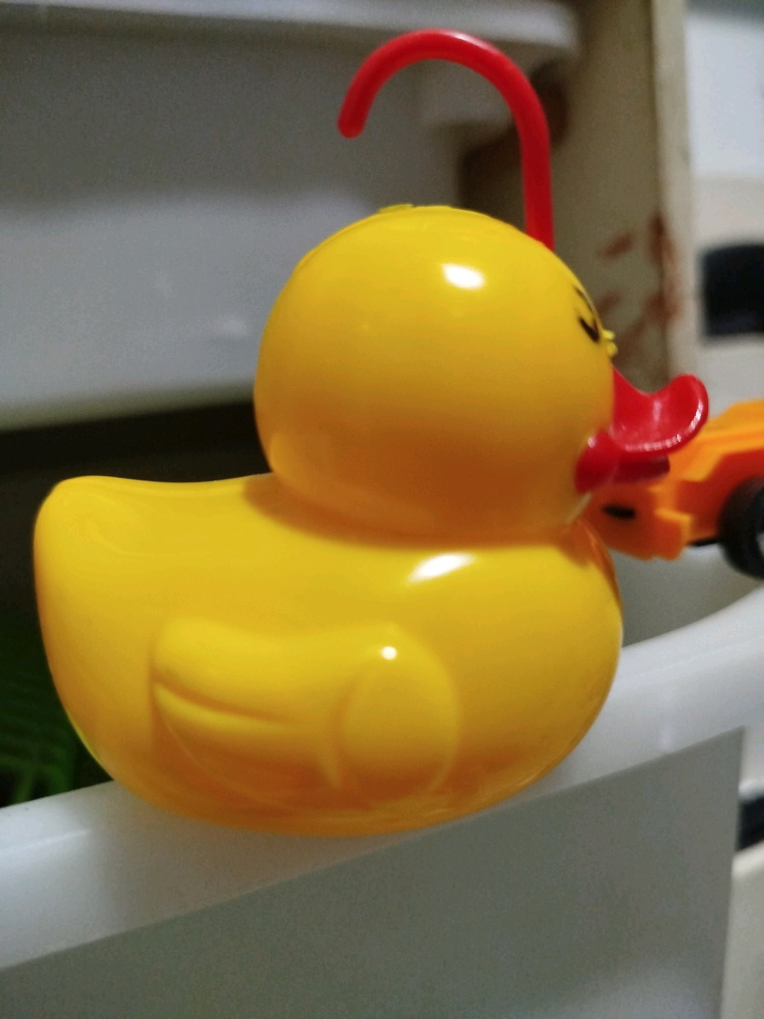 A bright yellow rubber duck toy with a red beak and a red loop is positioned on a white surface. The duck is facing to the right. In the background, a small orange toy car with black wheels is partially visible, attached to the duck's right side. The environment appears to be indoors, with a blurry background that suggests a domestic setting. There is no indication of a specific location within Mojoagung, Indonesia.