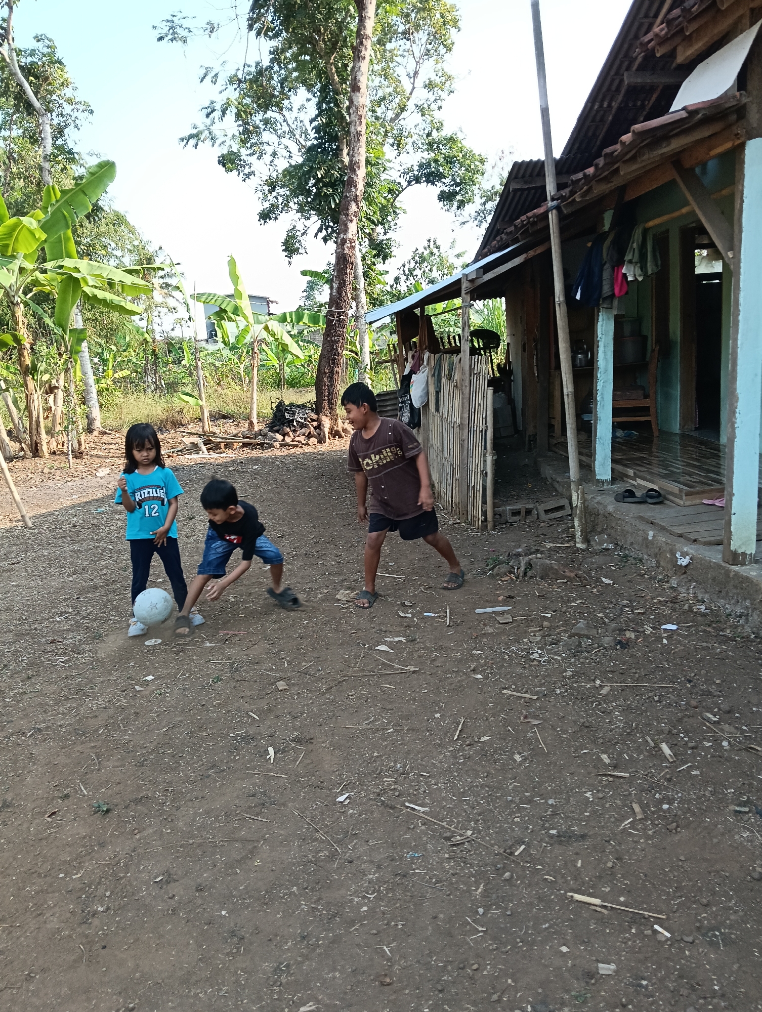 In Tasikmalaya, Indonesia, three children are playing with a soccer ball in a dirt yard in front of a modest wooden house. A young girl in a blue t-shirt with the number 12 and the word "RIZZLIE" on it stands with her hands on her hips, looking at the ball. A younger boy in a black t-shirt and blue shorts is crouched down, appearing to kick the ball. An older boy in a brown t-shirt and dark shorts stands to the side, watching. The house behind them is constructed with wooden posts and a corrugated metal roof, with clothes hanging on a line outside. Lush green banana trees and other vegetation surround the yard, indicating a rural or semi-rural setting. The ground is covered in dirt and scattered debris. The sky is bright and clear, suggesting it is daytime.