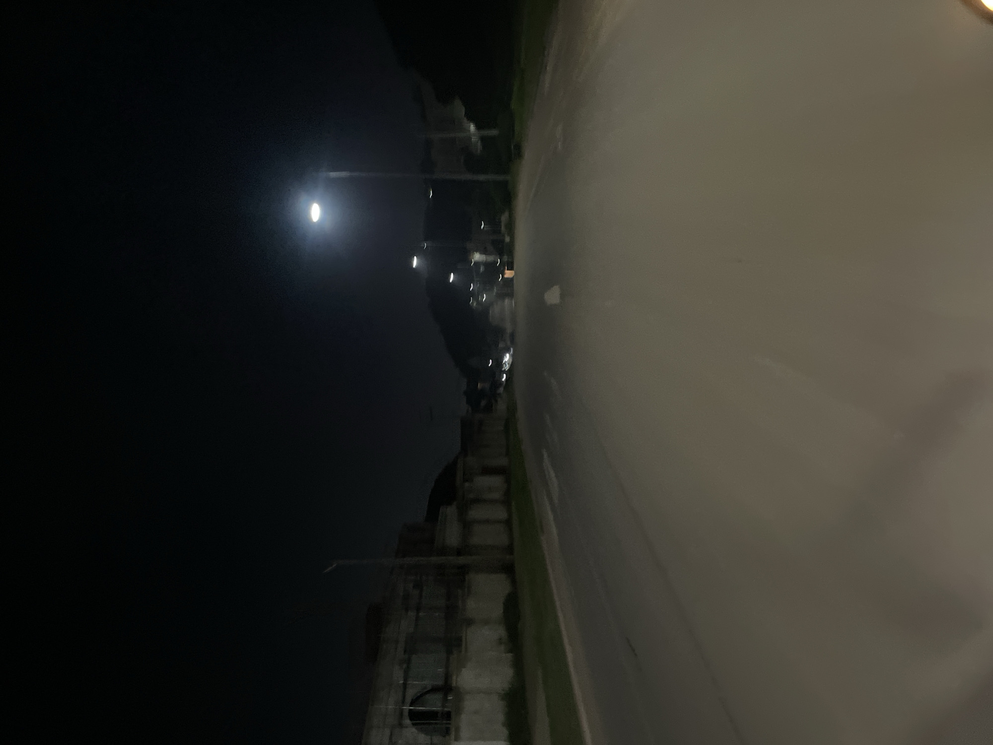 The image depicts a nighttime scene on an empty, paved road in Guzape, Nigeria. A street lamp illuminates the scene from the right side of the frame, casting a bright light on the asphalt. The road appears to be a straight, multi-lane stretch with faded white lane markings. On the left side of the road, there is a long, low-lying structure with a textured, light-colored facade and a darker roofline. Grass and some vegetation are visible between the road and the structure. In the distance, more street lights are visible along the road, and dark, silhouetted hills or buildings form the background under a dark night sky. There are no visible people or vehicles in the immediate vicinity. The overall impression is one of a quiet, dimly lit urban or suburban environment at night.