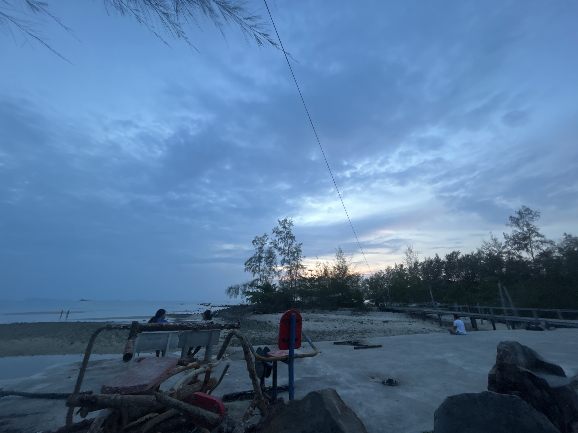 The image depicts a coastal scene in ตำบลเกาะพะงัน, Thailand, taken under a cloudy, pre-dawn or twilight sky. A beach is visible, with several indistinct figures present. One person is sitting on the beach near some vegetation, while two other figures are further away, walking in the shallow water. In the foreground, there are decaying exercise machines, consisting of a bench press and a seated exercise apparatus, indicating a public fitness area. A wooden walkway is partially visible leading into the trees on the right. Stones are also present in the foreground. A single electrical wire cuts across the upper portion of the image.