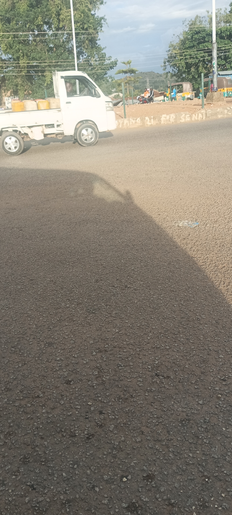 The image depicts a daytime street scene in Bukuru, Nigeria, observed from a low angle, likely from a stationary vehicle or structure casting a large shadow across the asphalt road.
In the midground, a white compact pickup truck, loaded with four yellow jerrycans in its bed, travels from left to right. A person, appearing to be an adult male, is visible in the driver's seat. To the right of the truck, further down the road, two individuals are seen on a motorcycle, and several auto-rickshaws (Keke Napep), primarily blue and yellow, are visible near the roadside.
The foreground consists of dark gray asphalt pavement, showing a coarse aggregate texture. A prominent dark shadow, originating from the lower left, extends diagonally towards the middle right of the frame, occupying roughly the left half of the road surface. A small piece of blue and white debris lies on the brighter, unshadowed part of the asphalt in the middle right.
The background features lush green foliage and trees lining the road. A white utility pole stands vertically on the right side, with overhead power lines extending across the upper portion of the frame. A low, light-colored wall runs along the right side of the road, on which partial white graffiti or painted text reads "YANKA DI FOR SENNE". The sky is partly cloudy with patches of blue visible at the top, and distant hills or landforms are discernible on the horizon.