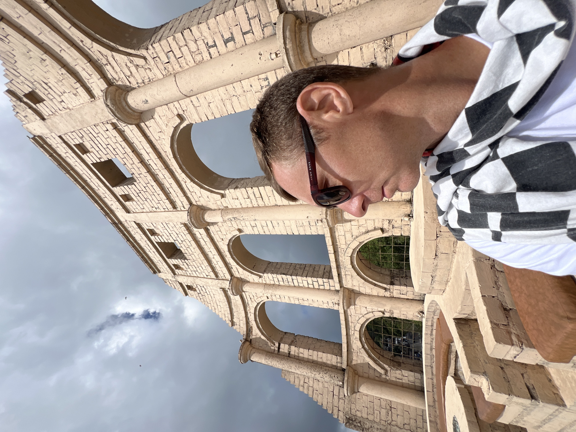 An outdoor, low-angle, medium shot captures a man in sunglasses and a checkered scarf standing in front of a large, beige, weathered stone structure resembling Roman architecture. The structure features arched openings and is partially ruined, with exposed brickwork. The sky is overcast and cloudy, with hints of blue visible through the clouds. The man is positioned on the right side of the frame, his profile facing left, and he is wearing a white t-shirt. The lower part of the image shows stone seating areas with brown cushions. This structure is part of the Kintamani area in Indonesia.
