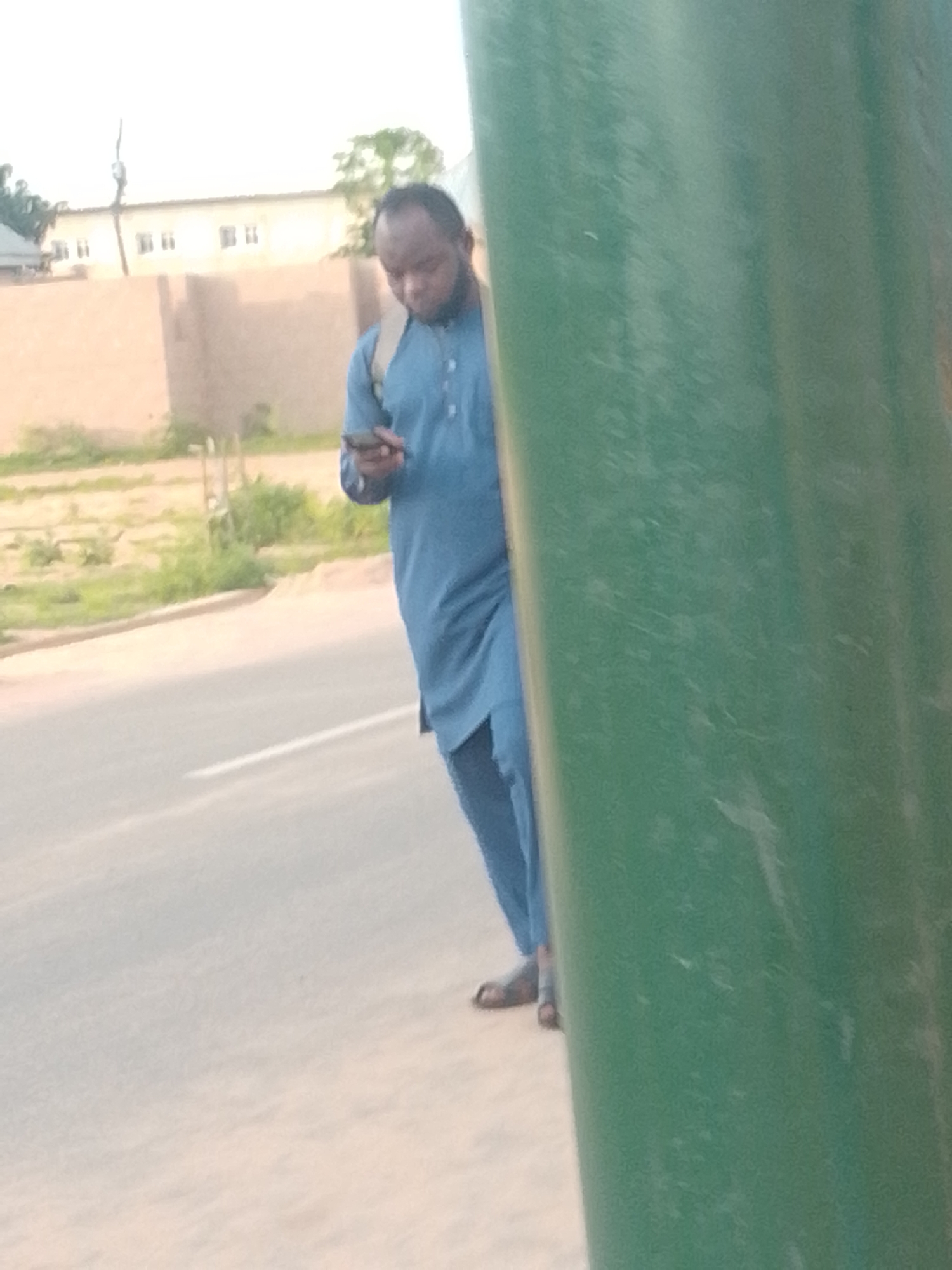 A man wearing a blue traditional outfit and sandals is standing on the side of a road, looking at his mobile phone. He has a backpack on his shoulders. The man is partially obscured by a large, green cylindrical object, possibly a pole or a pillar, on the right side of the frame. In the background, there are buildings and vegetation, suggesting an outdoor urban or semi-urban setting. The scene appears to be in Gombe, Nigeria, based on the context provided. The road is paved with a white line marking. The lighting indicates it is daytime.