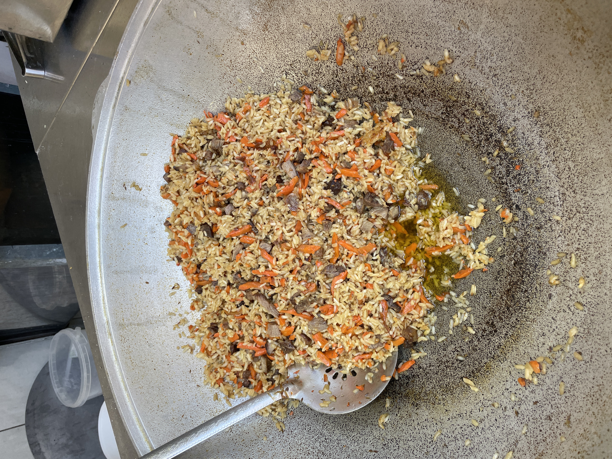 A close-up, overhead shot shows a large, round, metal pot filled with pilaf, a rice dish with carrots and meat. A metal slotted spoon rests on the left side of the pot, scooping some of the pilaf. The pilaf is a golden-brown color with visible chunks of orange carrots and dark meat. The pot itself appears to be made of cast iron or a similar material, with some oil and residue on the bottom. In the background, a stainless-steel counter and some plastic containers are visible, suggesting a commercial kitchen environment. The scene appears to be located in Sochi, Russia, likely within a restaurant or food preparation area.