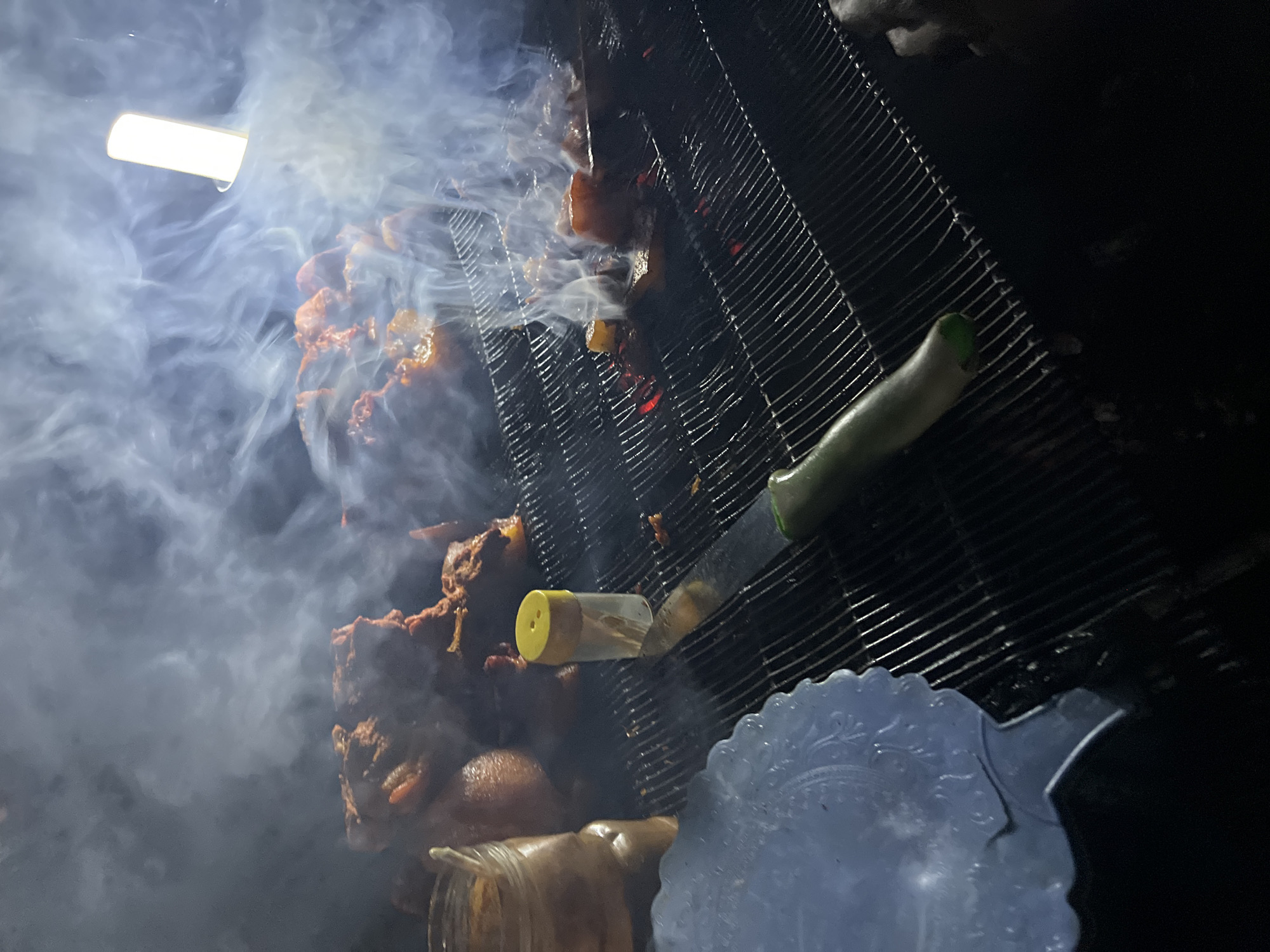 The image depicts a roadside food vending scene, possibly in Jos, Nigeria, showcasing skewered meat being grilled over hot coals. Dense smoke fills the foreground, partially obscuring the meat being cooked under a light source. A grilling rack is angled, with a knife with a green handle seemingly placed on the rack. Near the bottom of the image, there's a yellow-capped spice container, a jar, and a patterned plastic plate. The environment appears dark, lit by artificial lights. No individuals are visible.