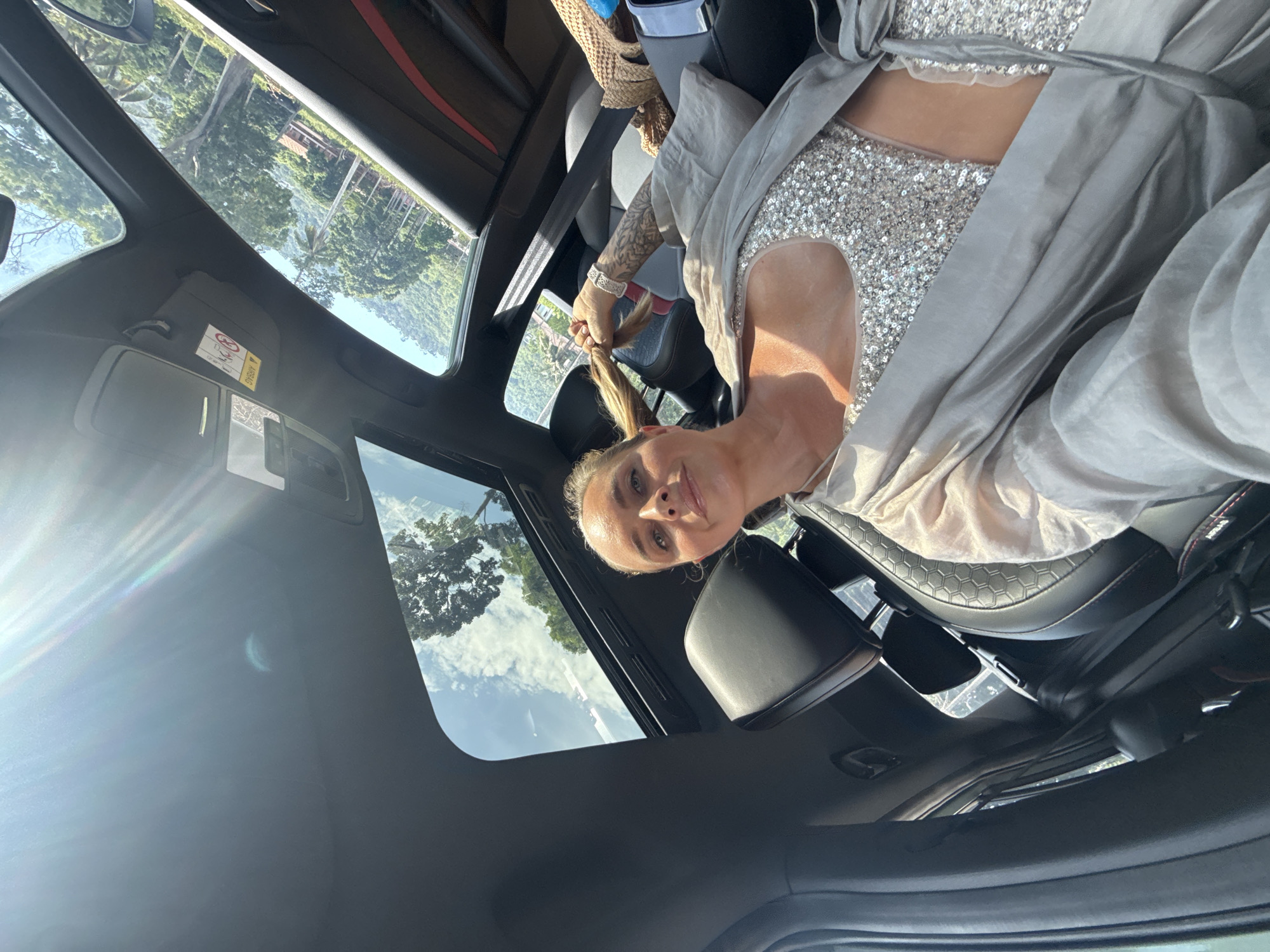 A woman is pictured in the passenger seat of a car, looking at the camera and holding a strand of her blonde hair. She is wearing a sequined top and a light gray robe. The car has a sunroof open, revealing a view of lush green trees and a blue sky with clouds. The setting appears to be a tropical location, possibly Ko Samui District, Thailand, given the vegetation and natural light. The interior of the car is visible, including black leather seats and a red accent trim. The overall mood is relaxed and sunny.
