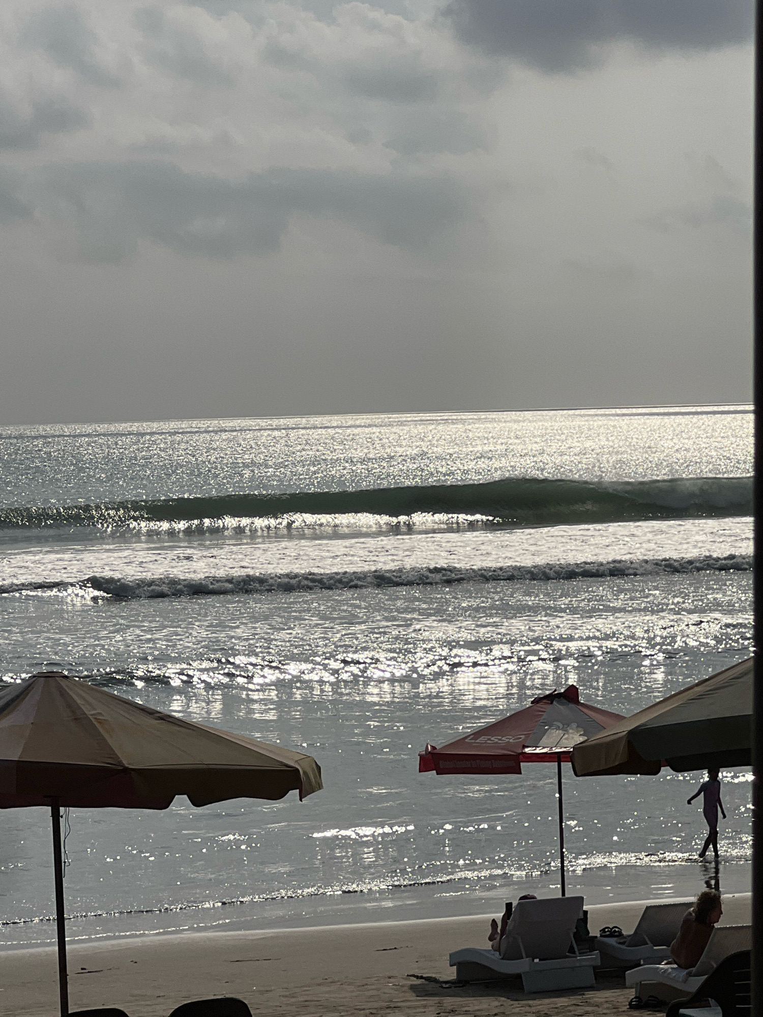 The image depicts a beach scene in Kuta, Indonesia, with the ocean in the background and umbrellas and lounge chairs in the foreground. The sun is shining brightly, casting a glittering reflection on the water and the wet sand. Several waves are rolling onto the shore, and a person is walking in the shallow water near the right side of the frame. A few lounge chairs are occupied by people relaxing on the beach. The sky is overcast with clouds, obscuring the direct sunlight and creating a diffused light across the scene.