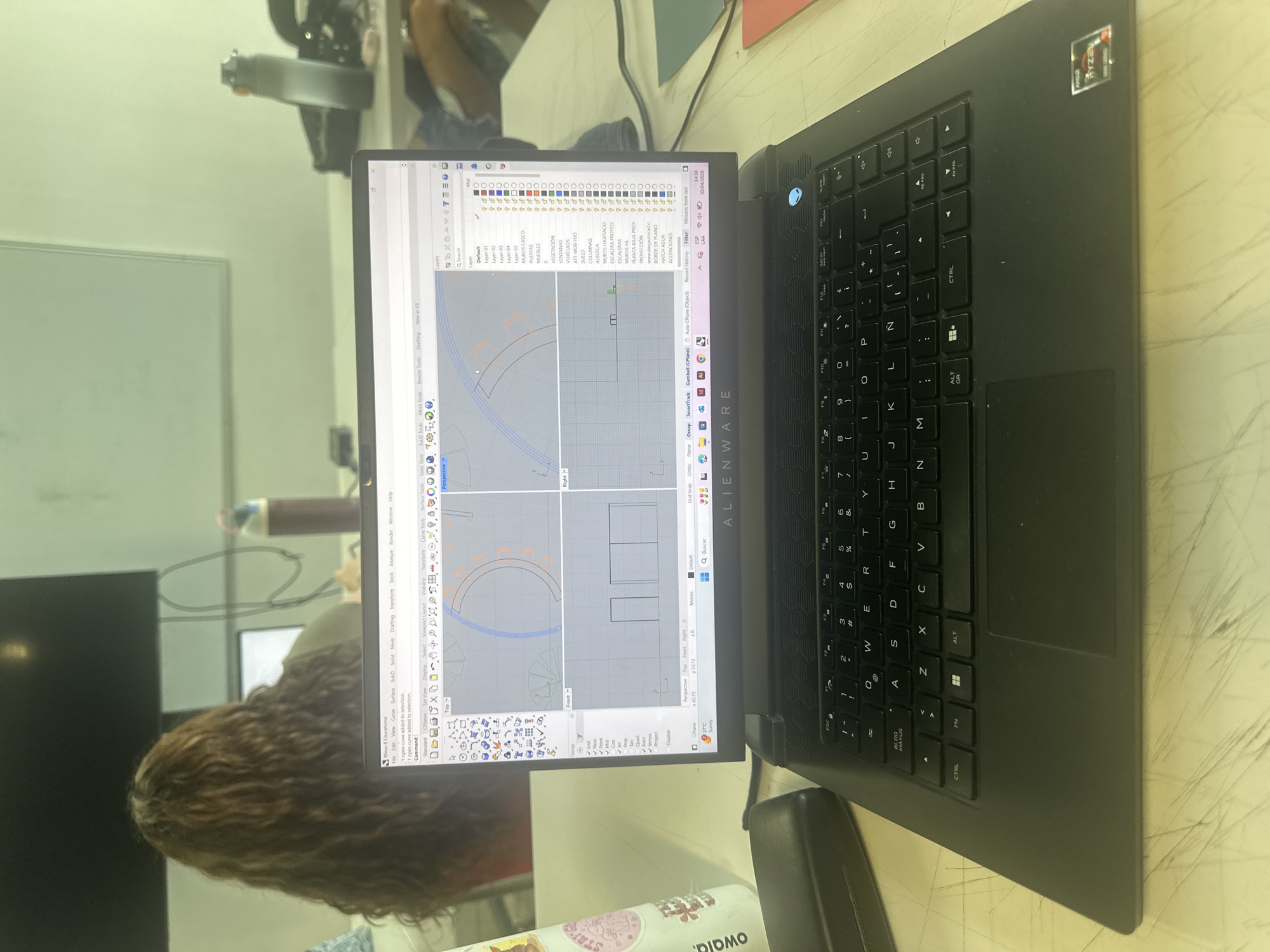 An Alienware laptop with an AMD Ryzen processor is displayed on a light-colored desk, showing a CAD software interface (Rhino 8 Educational). The laptop is black with a backlit keyboard featuring a QWERTY layout with Spanish characters. On the screen, a 2D architectural or engineering design is visible, segmented into multiple views (Perspective, Top, Front, Right). The software's layer panel shows various elements like "MUROS CASCO" (hull walls), "PUERTAS" (doors), "MUEBLES" (furniture), "VEHÍCULOS" (vehicles), "COLUMNAS" (columns), "ESCALERAS" (stairs), and "ACOTACIONES" (dimensions), indicating a detailed building plan.
To the left of the laptop, the head and curly brown hair of a person are partially visible, suggesting they are actively using the computer. A white and pink Owala brand water bottle, with "STAYE" text visible on a sticker, sits on the desk near the laptop. In the background, a white board and part of a chair are faintly discernible, hinting at an indoor setting such as an office, classroom, or study area. The laptop screen also displays the date "30/04/2026" and weather information "27°C Sunny". The scene suggests a daytime activity focused on design or drafting work.