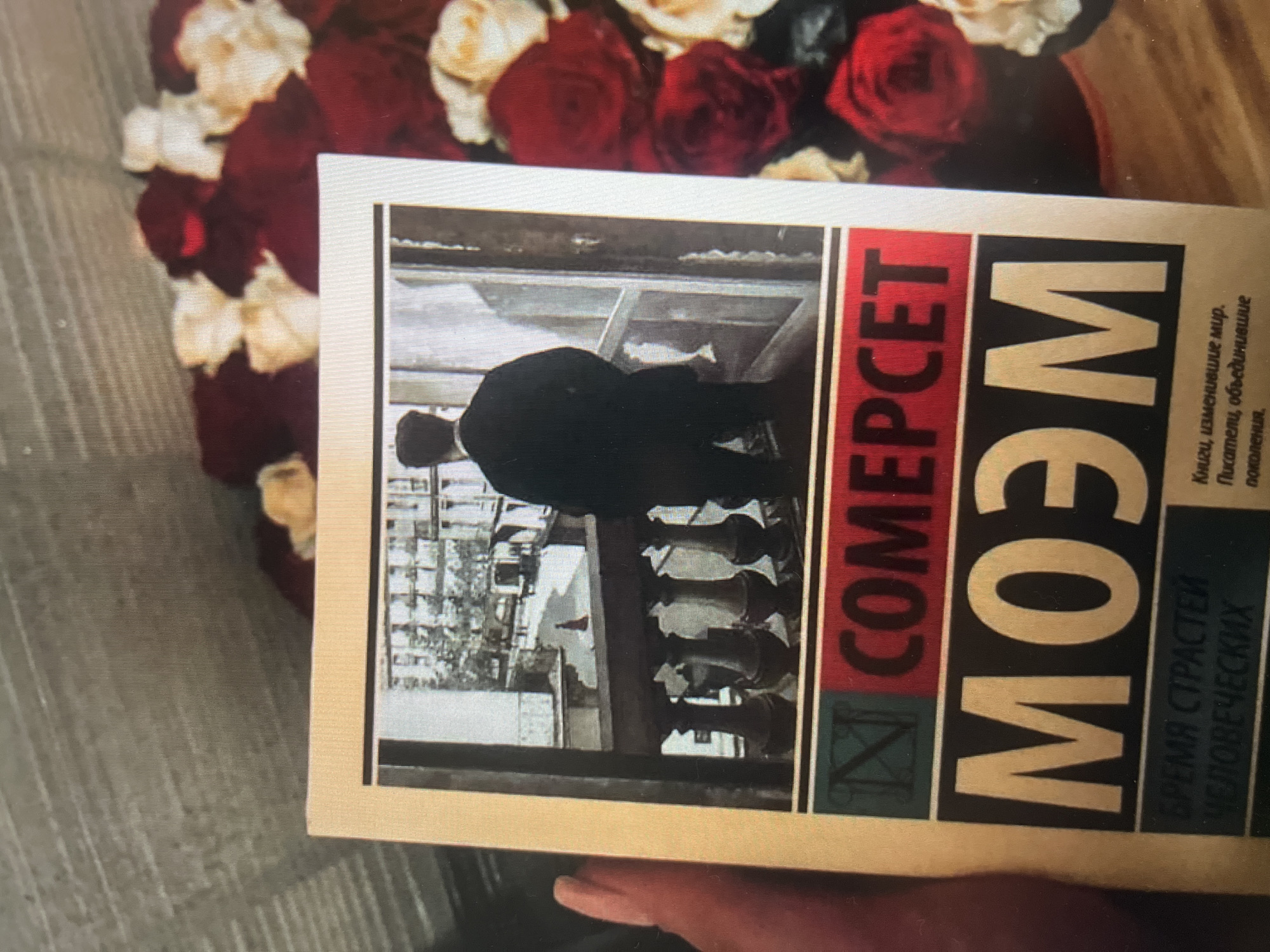 A book cover, likely a Russian edition, is held in a person's hand. The book features a black and white image of a man in a suit standing on a balcony, looking out over a city street. The text on the cover is in Russian. Below the image, the title reads "МОЭМ" in large white letters on a black background. Above this, the word "СОМЕРСЕТ" is printed in red letters on a dark green background. Smaller text at the bottom of the cover includes "ВРЕМЯ СТРАСТЕЙ ЧЕЛОВЕЧЕСКИХ" and "Книги, изменившие мир. Писатели, объединившие поколения." The book is positioned in front of a bouquet of red and white roses. The background is blurred but appears to be a textured surface, possibly a wall or fabric.
