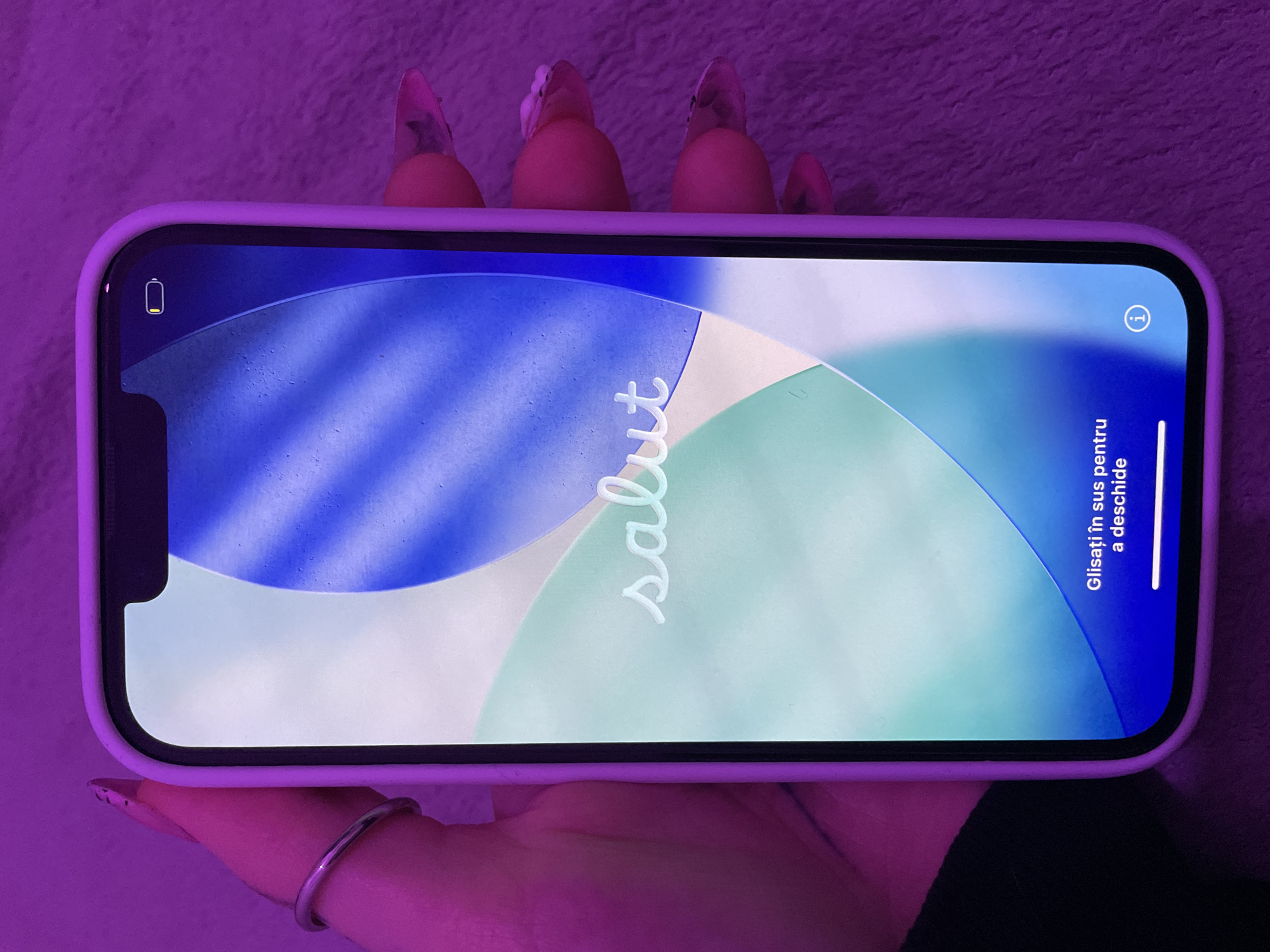 A hand holds an iPhone displaying a locked screen. The screen reads "Salut" in cursive, with "Glisați în sus pentru a deschide" below, indicating the phone is set to Romanian. The battery icon in the top left corner shows a low charge. The phone is in a lilac-colored case. The background is a soft, blurry purple texture.
There is no information on the location of the city of Buenos Aires, Argentina in the provided image.