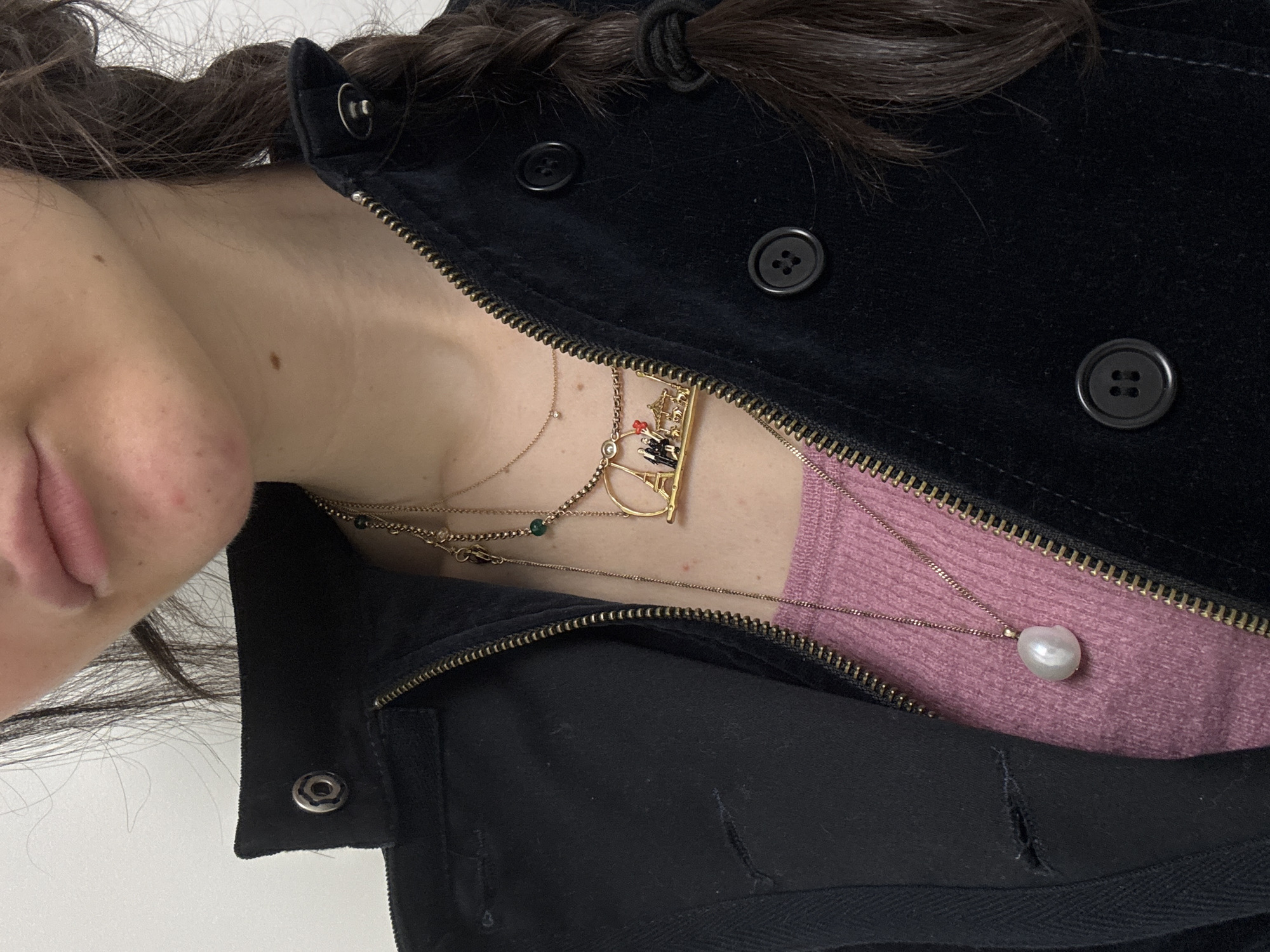 The scene depicts a close-up, chest-level view of an unidentified individual in the city of New York, United States of America, against a stark white background. The subject is dressed in a dark, possibly corduroy or suede-like, collared jacket, partially unzipped, revealing a ribbed pink garment underneath. The jacket features a metal snap on the left collar and multiple dark buttons on the right lapel. Several gold-toned necklaces are layered around the neck. These include a delicate chain with a small round pendant, a chain with a green bead and a red charm, a prominent charm depicting the Eiffel Tower with two figures and a red balloon, and a longer chain with a large, white, irregular baroque pearl pendant. The lower portion of the subject's face, including the chin and lips, is visible, along with a mole on the right side of the neck and minor facial blemishes. Dark brown hair, styled in a braid, extends over the right shoulder and is secured with a black hair tie.