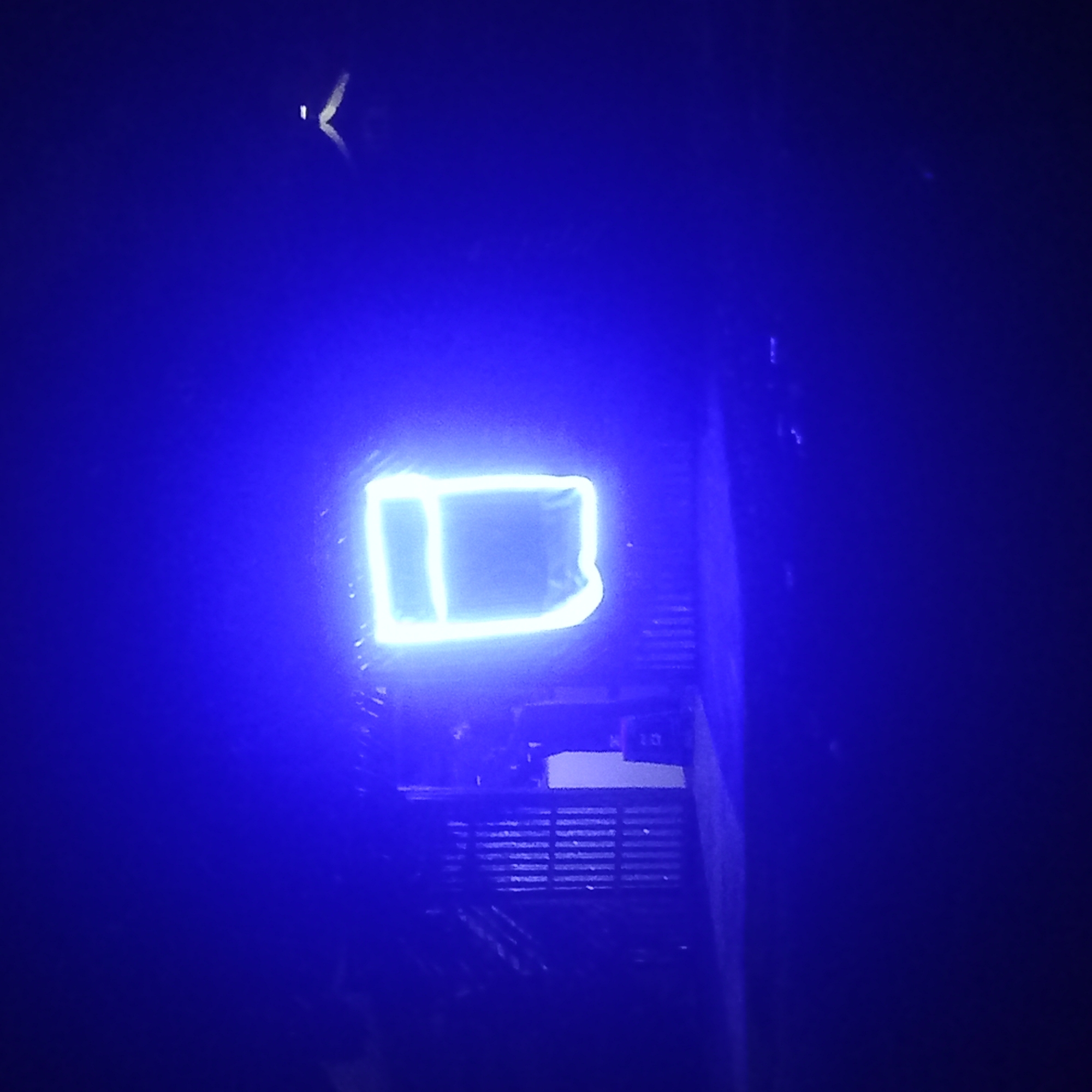 A luminous, blue, stylized letter "D" is the primary subject of the image. It appears to be a neon or LED light. The letter is positioned against a dark background, appearing to be inside or in front of a structure with horizontal grill-like features. Elements of electronic equipment or components are also visible, partially obscured by the darkness. The overall scene is very dark and dimly lit, suggesting a nighttime setting. The location and city of Barden Yana, Nigeria are not evident in the imagery.