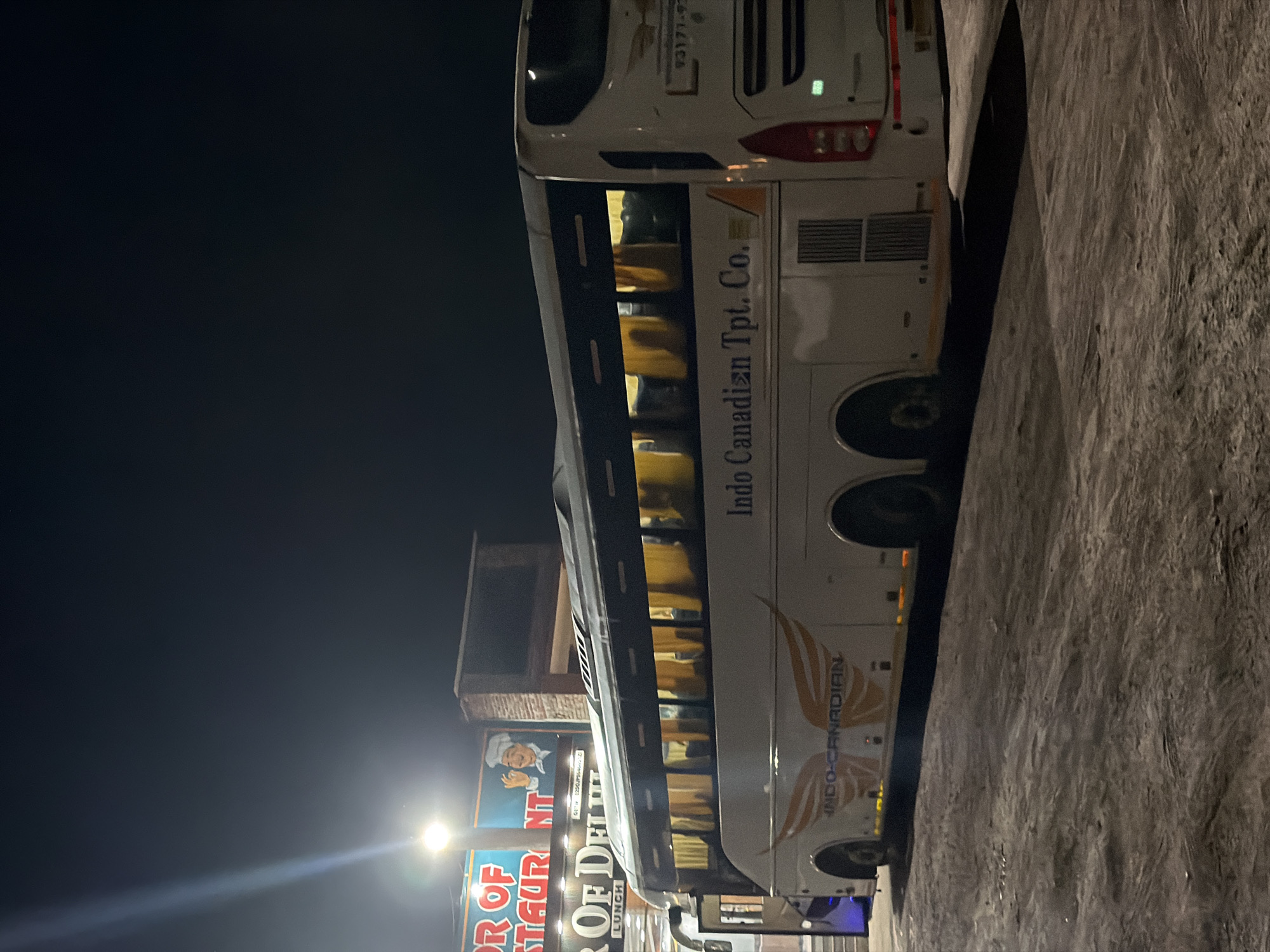 A white bus with "Indo Canadian Tpt. Co." written on its side is parked at night next to a textured, grey surface, possibly a wall or embankment. The bus has illuminated windows, revealing rows of seats inside. A sign for "OR OF RESTAURANT" and "OF DELHI LUNCH" is visible to the left of the bus. Another sign with "INDO-CANADIAN" and a stylized wing graphic is present on the bus. The lighting suggests it is nighttime, with a bright light source to the left casting a glare. The scene appears to be in a commercial or travel-related area, given the restaurant signage and the bus. There are no people clearly visible in the image. The overall atmosphere is dark and somewhat utilitarian.