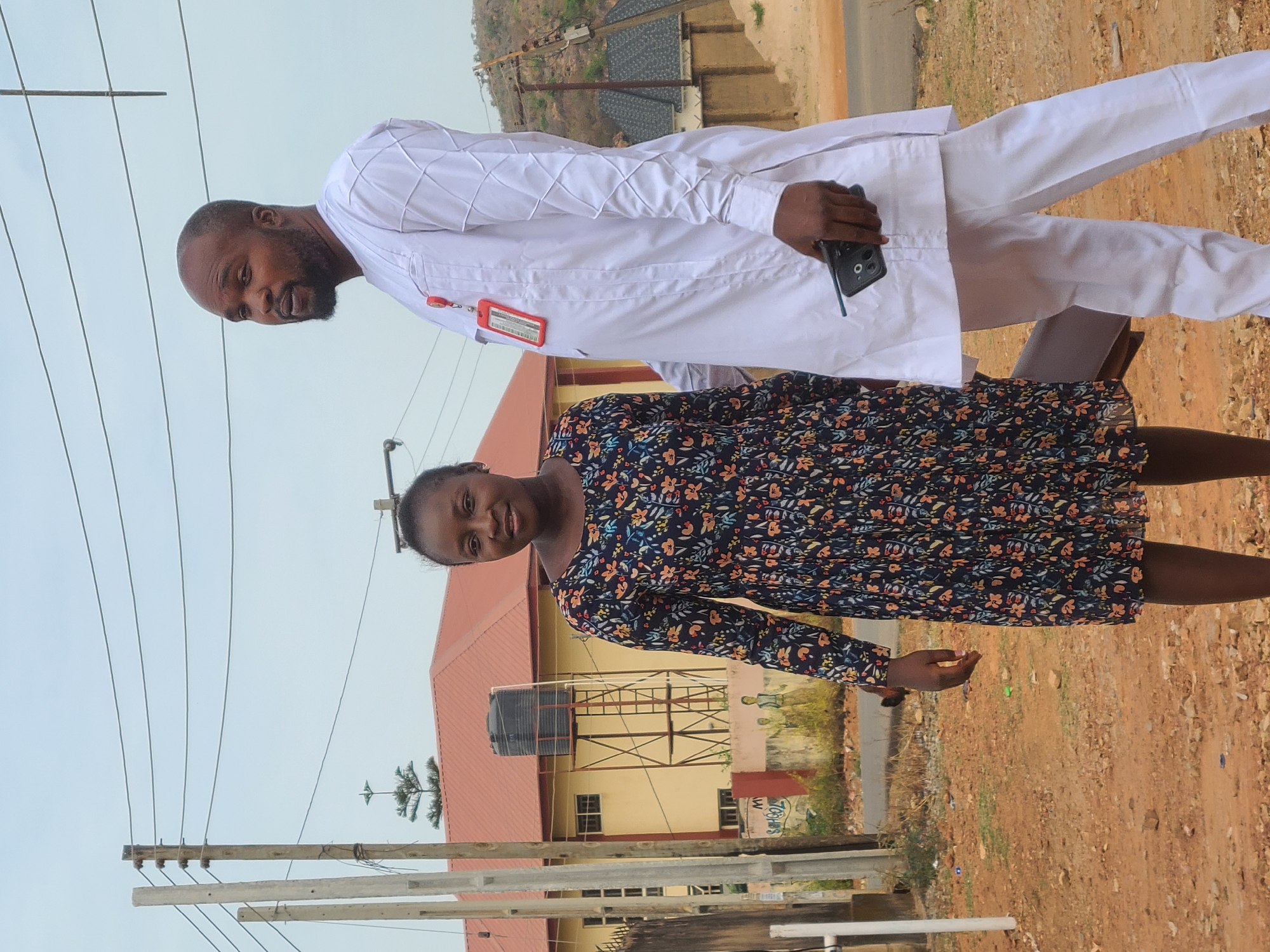 The image shows two people standing outdoors. A man with a beard, wearing a white, long-sleeved outfit with white pants and holding a smartphone, is positioned to the left. Next to him, a woman with her hair pulled back is wearing a dark, floral-patterned dress. They are both facing the camera. Behind them is a building with a red roof and a water tank on a steel frame. Utility poles and power lines are visible. The ground is dirt and gravel. The scene is Gura Top, Nigeria.