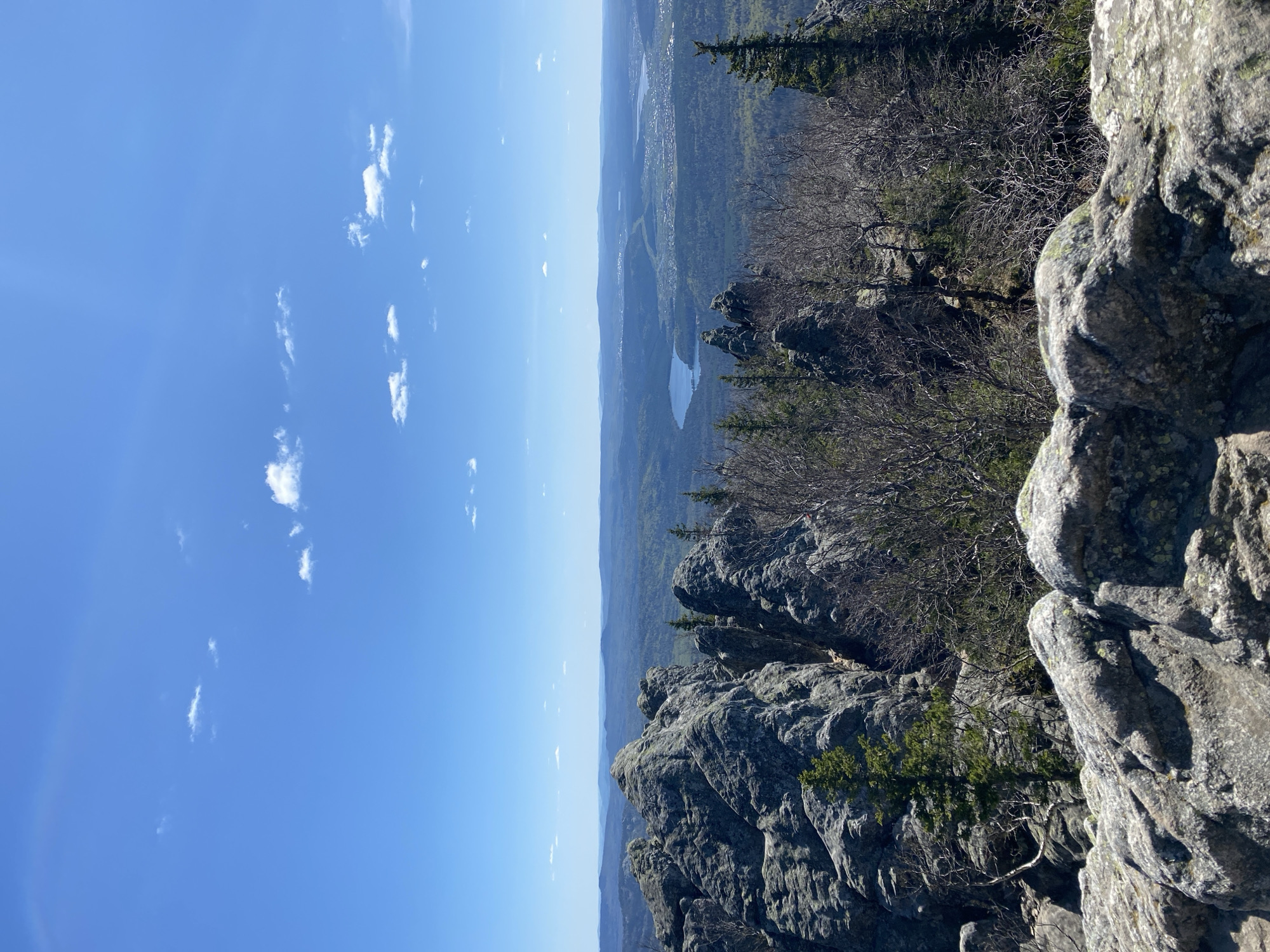 The image shows a rocky overlook with a view of the city of Zlatoust, Russia. The foreground is dominated by large, gray rocks, some with small trees growing on them. The background shows a valley with a lake and the city in the distance. The sky is blue with scattered white clouds.