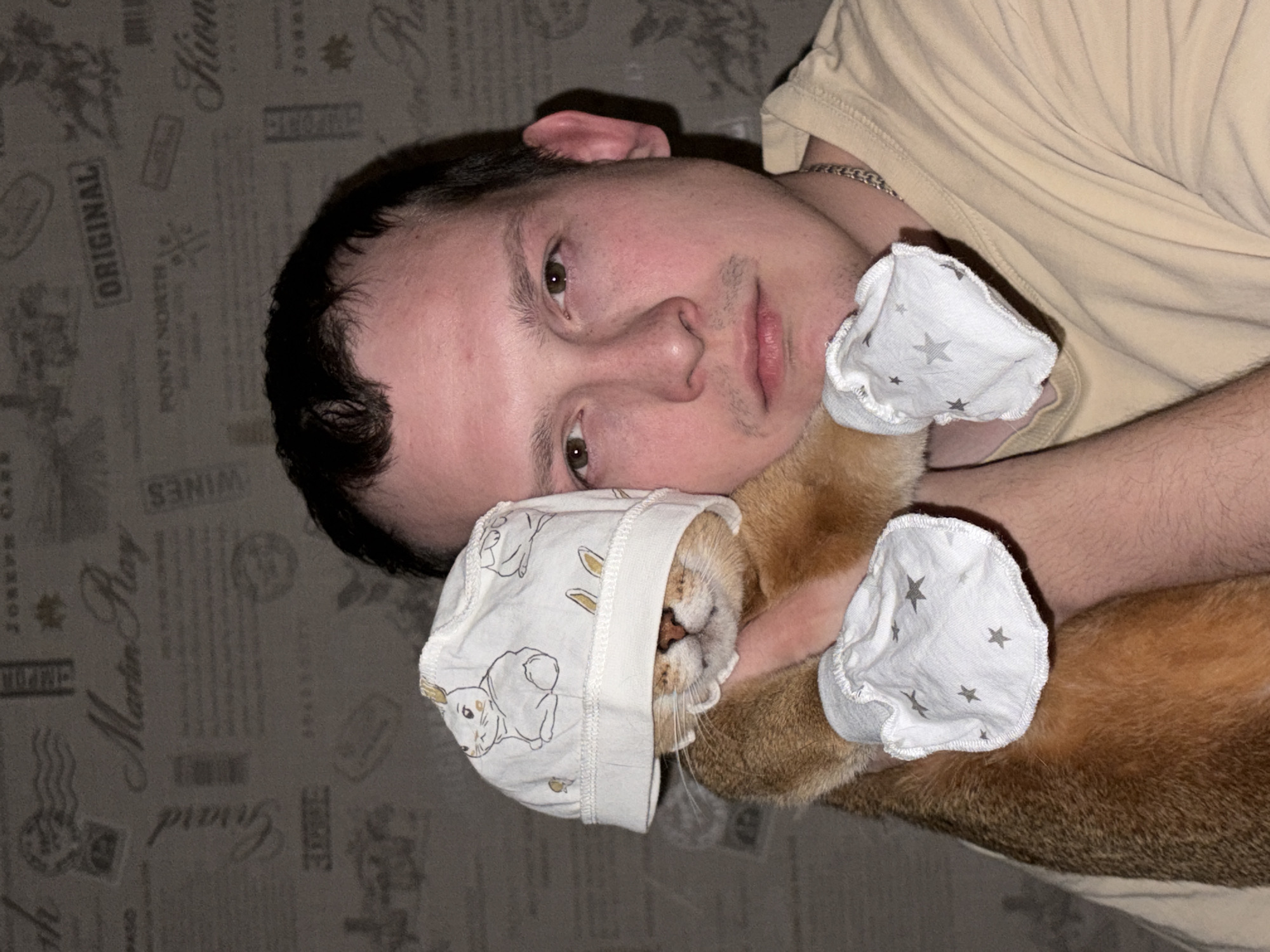 In an indoor setting in Chaikovsky, Russia, a male individual is depicted holding a domestic cat. The male, appearing to be between 20 and 30 years old, has short dark hair, light skin, and faint stubble on his chin and upper lip. He wears a light-colored, short-sleeved t-shirt and a silver-toned chain necklace. He looks directly at the camera with a neutral expression. The cat, with short, reddish-brown fur, is positioned against the man's chest/shoulder and is wearing a white fabric cap patterned with rabbits, which covers its eyes and ears. Its front paws are covered by white fabric mitts adorned with gray stars. The man's right arm is visible, supporting the cat. The background is a wall covered with patterned wallpaper displaying faded text and designs, including words such as "Martin Ray," "Girard," "Joseph Care," "Point North," "Original," and "Wines."