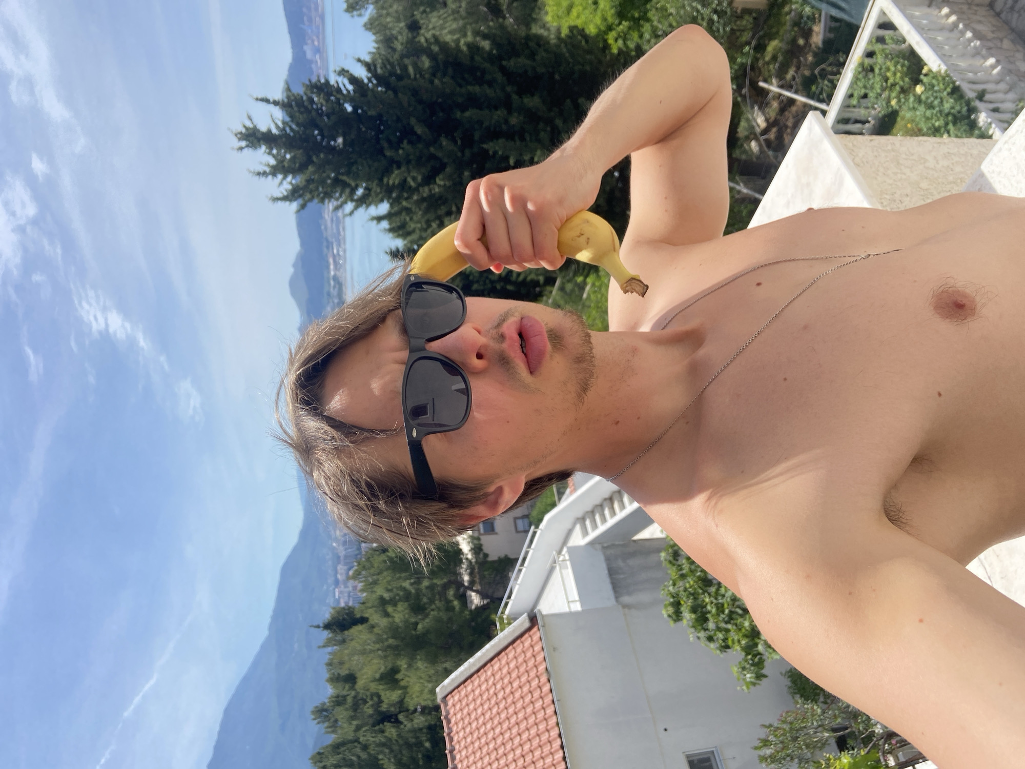 A man is holding a banana to his ear, wearing sunglasses, and shirtless. He has a thin silver chain around his neck. In the background are mountains, trees, buildings, a blue sky, and water. The photo was taken in Šušanj, Montenegro.