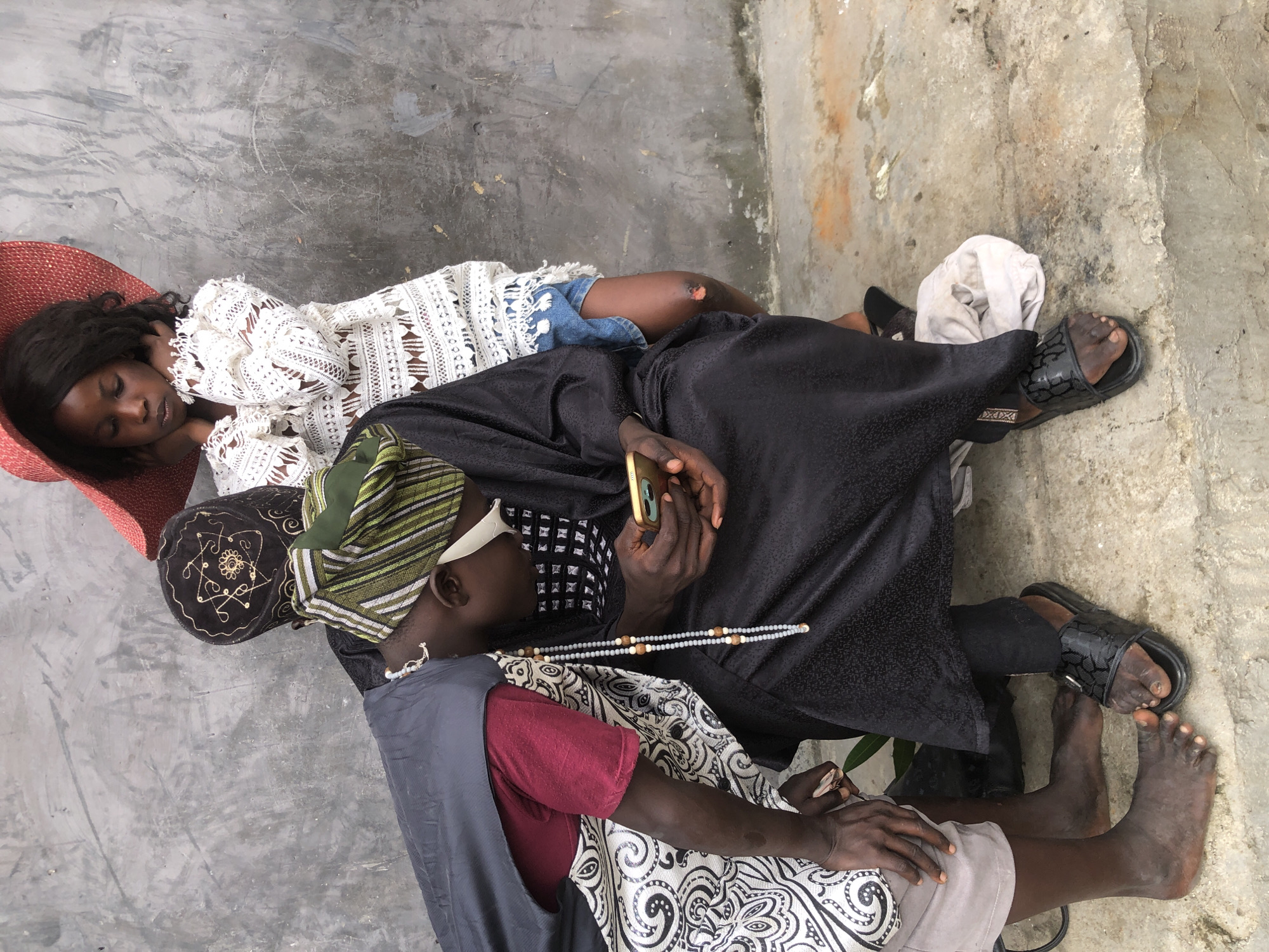 In Ado, Nigeria, three individuals are positioned on a concrete surface against a textured gray wall. In the foreground, a person wearing a dark robe and a green and white striped cap is seated, engrossed in their mobile phone. They have a string of light-colored beads around their neck. To their left, another person, clad in a grey vest over a maroon t-shirt and a patterned white and black skirt, leans forward. Their bare feet are visible. Behind the seated person, a young girl stands, wearing a large, red hat and a white lacy garment, with denim shorts underneath. Her hands are clasped near her neck. The ground is a rough, light-colored concrete with some dirt and debris.