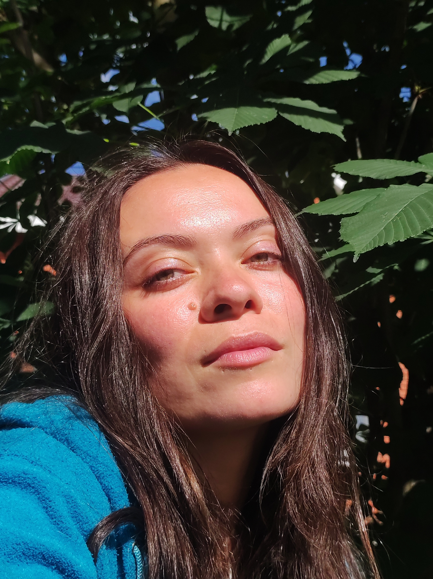 A close-up, overhead shot captures a young woman's face, angled slightly upwards. Her long, dark brown hair is loose, framing her face and falling over her shoulders. She wears a bright blue hooded jacket. Her skin is illuminated by sunlight, creating a warm glow with some pinkish undertones and highlighting a small mole on her cheek. Her eyes are looking slightly off-camera, and her lips are slightly parted. Behind her, lush green foliage from trees creates a natural backdrop, with dappled sunlight filtering through the leaves, suggesting an outdoor setting. The overall mood is serene and natural, hinting at a sunny day. There is no visible text in the image.