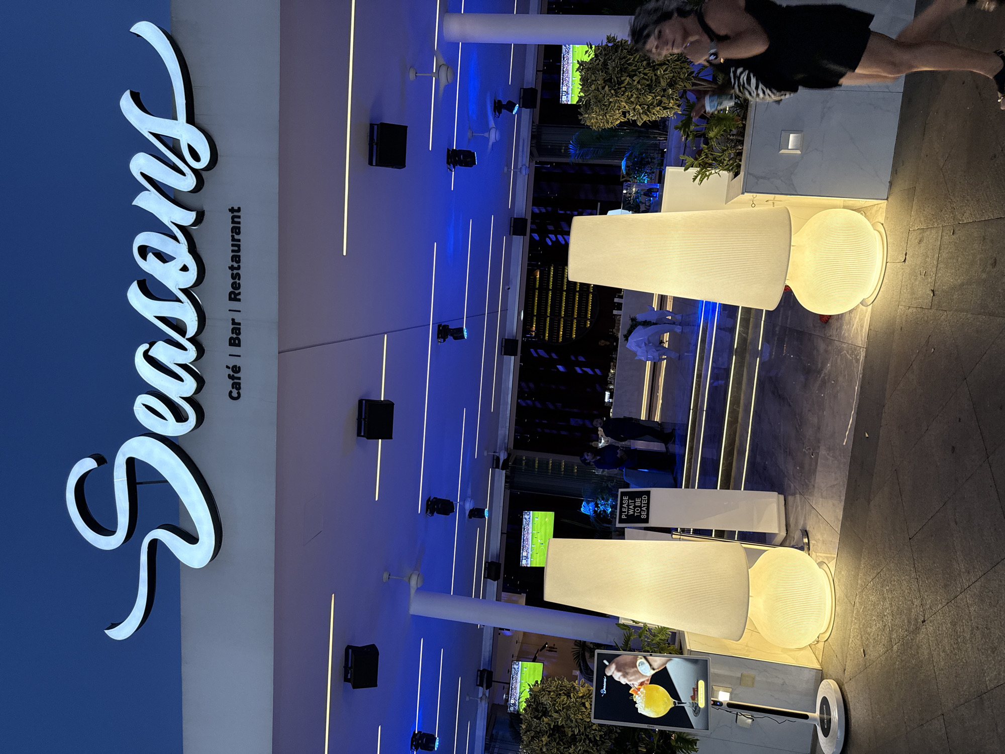 The image depicts the entrance of a venue named "Seasons Cafe | Bar | Restaurant" in Ayia Napa, Cyprus, at dusk. The prominent signage features stylized white lettering against a deep blue sky. The entrance is modern, with a white facade, recessed lighting, and blue ambient lighting overhead. Large, textured lamps illuminate the pathway leading into the venue. Two large screens displaying a football match are visible inside. Several individuals are present: a woman in the foreground walks from right to left, holding a cup and wearing a black dress and sandals. Two figures stand near a "Please Wait To Be Seated" sign, and other patrons are visible within the establishment. The overall atmosphere suggests a contemporary dining and entertainment establishment.