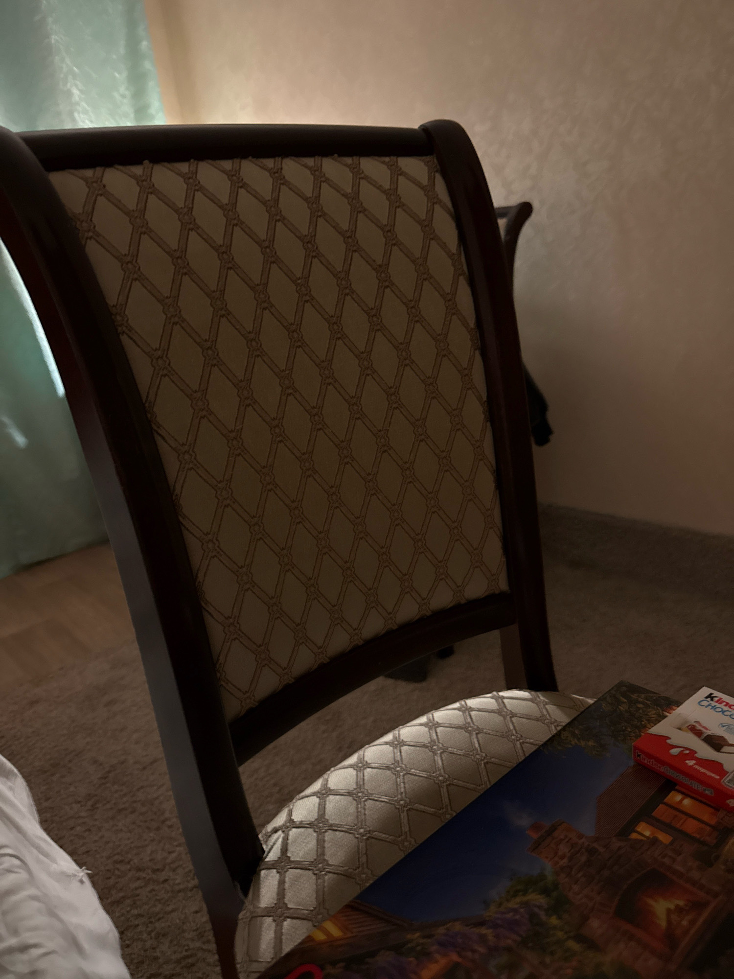 The image depicts an interior scene featuring a dark wooden armchair with patterned upholstery. The chair's backrest and seat are visible, showcasing a diamond-shaped, possibly damask, pattern in a light neutral color. A portion of a white sheet or blanket is draped over the left side of the chair. In the foreground, resting on the seat of the chair, is a book or magazine with an image of a cozy scene featuring a fireplace and a brick house. Next to it is a red and white Kinder Chocolate box. The background shows a light-colored wall and a glimpse of a window with a sheer curtain. The lighting is dim, suggesting it might be evening or a low-light interior. There are no people visible in the image. The location is not specified in the image itself, and there is no information linking it to Zavolzhye, Russia.