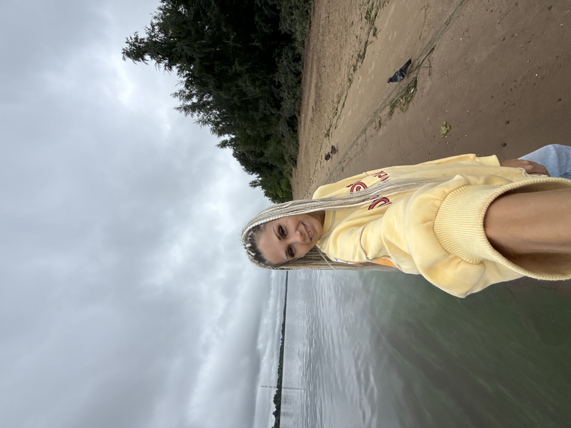 A woman with blonde braided hair is taking a selfie outdoors by a body of water. She is wearing a yellow sweatshirt and jeans. The water is calm and a beach with trees lines the background. Two birds are visible on the sand. The sky is overcast and cloudy.