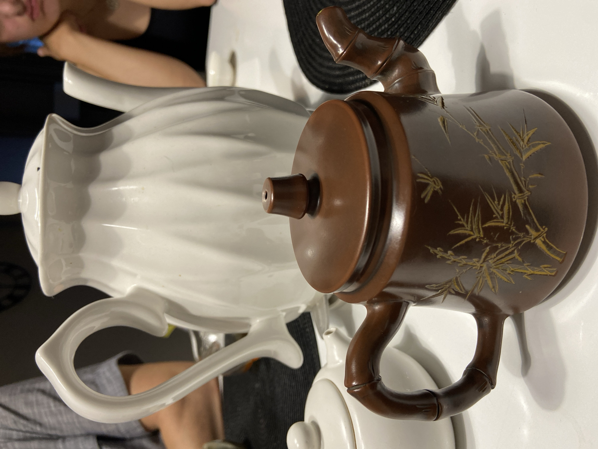 The image depicts a close-up view of two teapots on a white table. In the foreground, a small, dark brown teapot with a bamboo-shaped handle and a lid is prominent. The teapot is adorned with a golden bamboo leaf design. Behind it, a larger white teapot with a fluted design and a curved handle is partially visible. To the left, a smaller white teapot with a round lid is also in the frame. In the background, a person wearing a grey t-shirt is seated on the left, and another person in a black top is visible on the right, leaning their head on their hand. The setting appears to be an indoor environment, possibly a dining area. The image does not contain any information that would allow for its location to be determined in Blagoveshchensk, Russia.