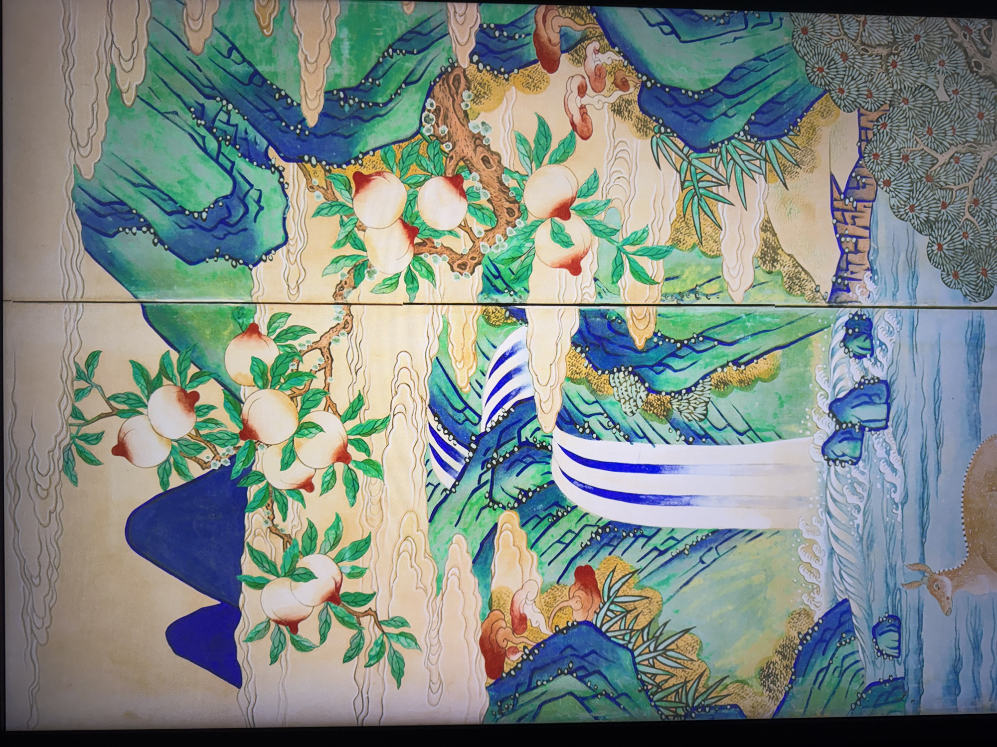 This is a close-up shot of a vibrant, two-panel folding screen painting. The painting depicts a stylized landscape with a waterfall, mountains, and fruit trees. In the foreground of the left panel, a tan deer with spotted markings stands in calm, light blue water. The waterfall cascades down from the upper portion of the screen, with blue and white stripes indicating flowing water. The surrounding landscape features lush green mountains with blue accents, rendered in a somewhat abstract, almost geological style. Wisps of pale yellow clouds drift across the scene.
On the upper branches of a tree on the left side of the screen, several ripe, pale pink peaches with red blush are depicted, surrounded by green leaves. Similar peach branches are visible on the right panel. The right side of the screen continues the mountainous landscape, with more green and blue formations. In the middle ground of the right panel, a cluster of red and brown mushrooms grows from the rocky terrain. The bottom right corner of the screen features a dense cluster of stylized evergreen trees with needle-like foliage. The overall style is reminiscent of traditional East Asian art, with bold colors and simplified forms. The painting appears to be on a fabric or silk surface, with a visible texture. The image is a photograph of the artwork.