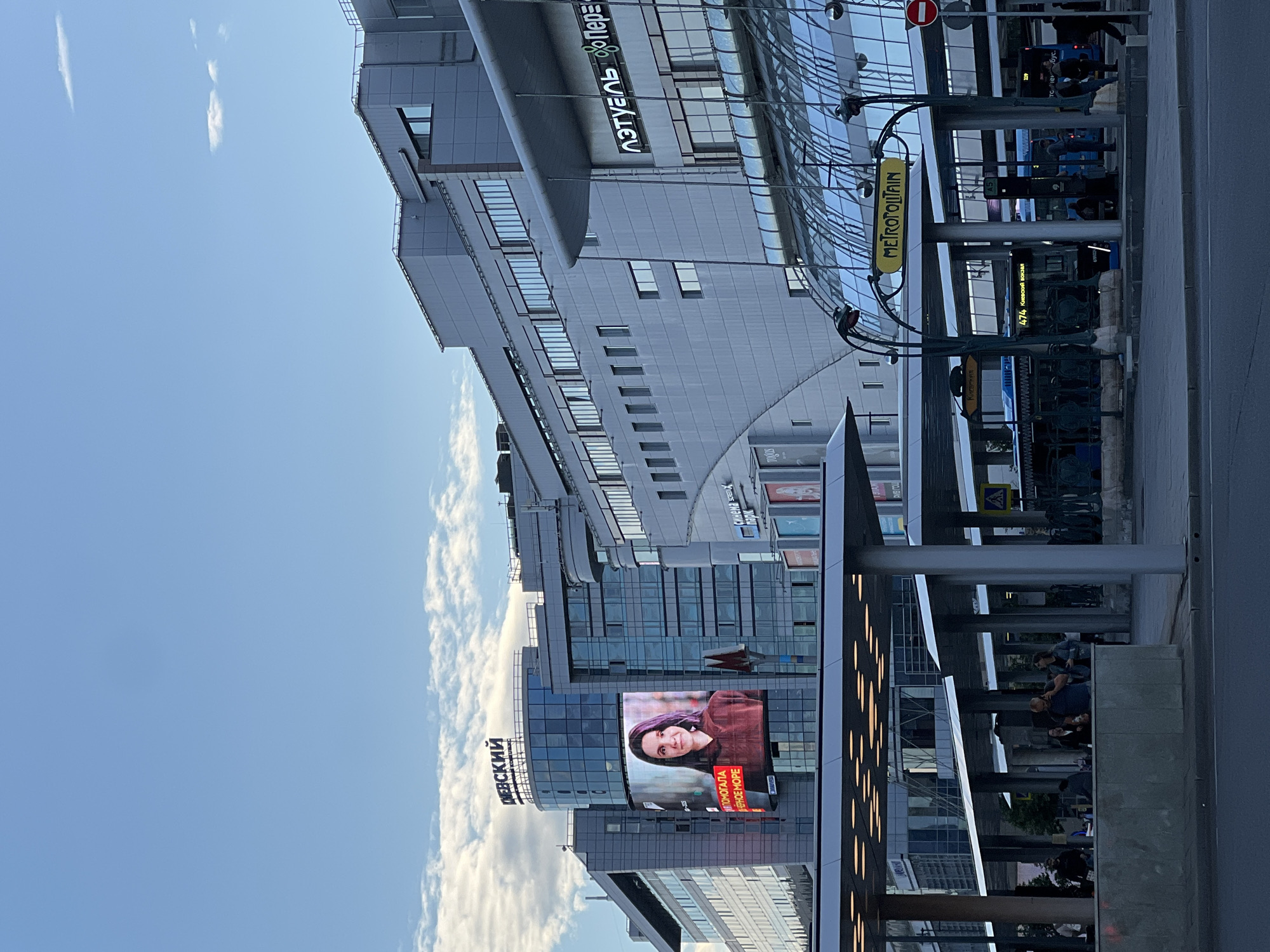 This image, taken in Moscow, Russia, depicts a street scene with a modern architectural setting. The composition is dominated by a large, multi-story building with a glass facade, reflecting the sky and clouds. In the foreground, there is a covered walkway or bus stop area with several pillars and people gathered. A large digital billboard displays a portrait of a woman and Russian text.
A prominent sign with the word "METROPOLITAIN" in yellow lettering on a blue background suggests the proximity of a subway station. Other visible text includes "ДЕВСКИЙ," "МЕПОМОГАЛА," "EPHOE MOPE," "синема парк," and "пэтуль Пере," which likely refer to businesses or advertisements within the area. The sky is a clear blue with scattered white clouds, indicating daytime. The presence of people waiting and walking suggests daily urban activity. The lighting suggests it might be late afternoon or early evening, given the shadows and the illumination of the signs. The overall atmosphere is one of urban commerce and transit.