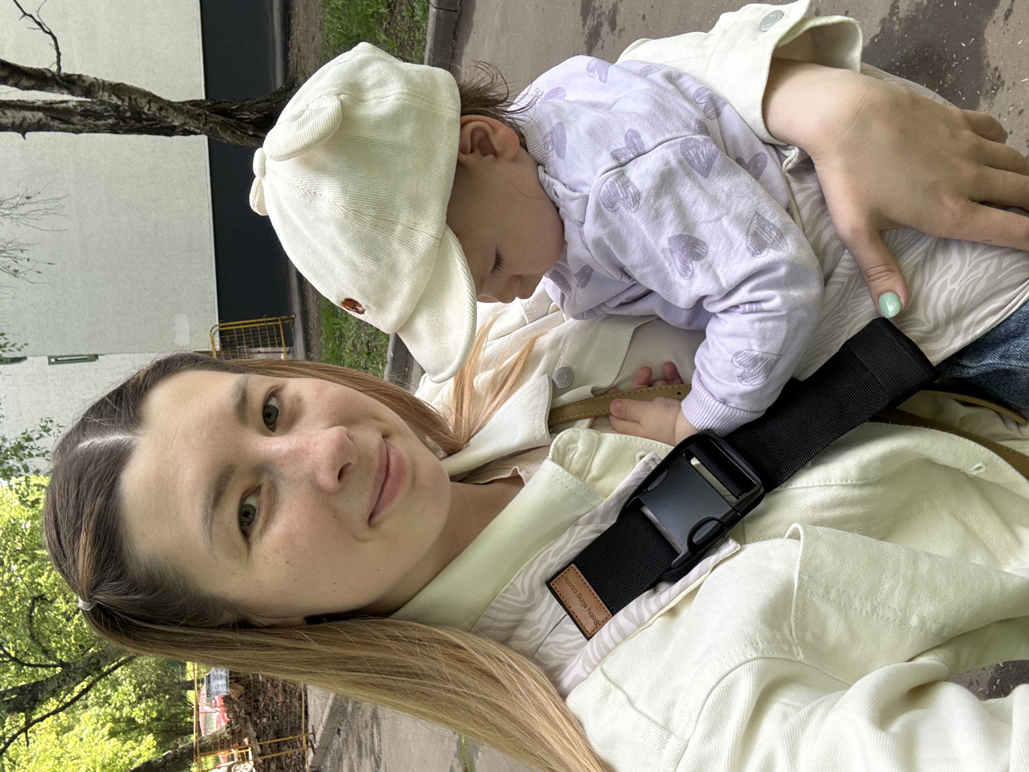 A woman with fair skin and long blonde hair is holding a baby in a carrier. The baby is wearing a light purple sweatshirt with heart patterns and a white cap. The woman is smiling and looking at the camera. A black buckle strap is visible on the carrier. The background shows a street with trees and buildings, with a partial view of birch tree bark. The photo was taken in the city of Moscow, Russia.