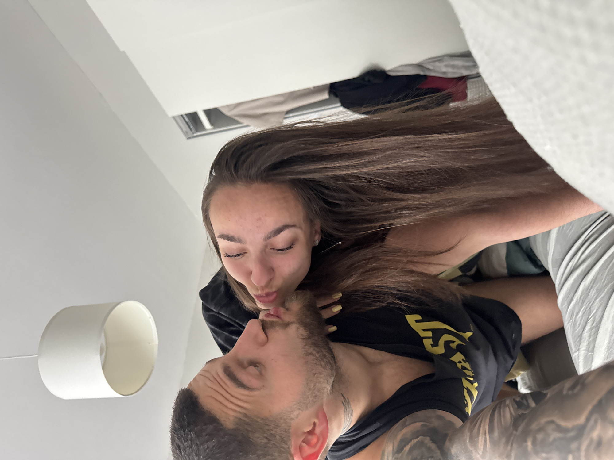 A woman with long brown hair and a man with short hair are kissing. The woman has her eyes closed. Both are wearing black t-shirts. The man has tattoos on his neck and arm. They are inside, near a bed and a lamp. The location is indeterminate, but the city is Sofia, Bulgaria.