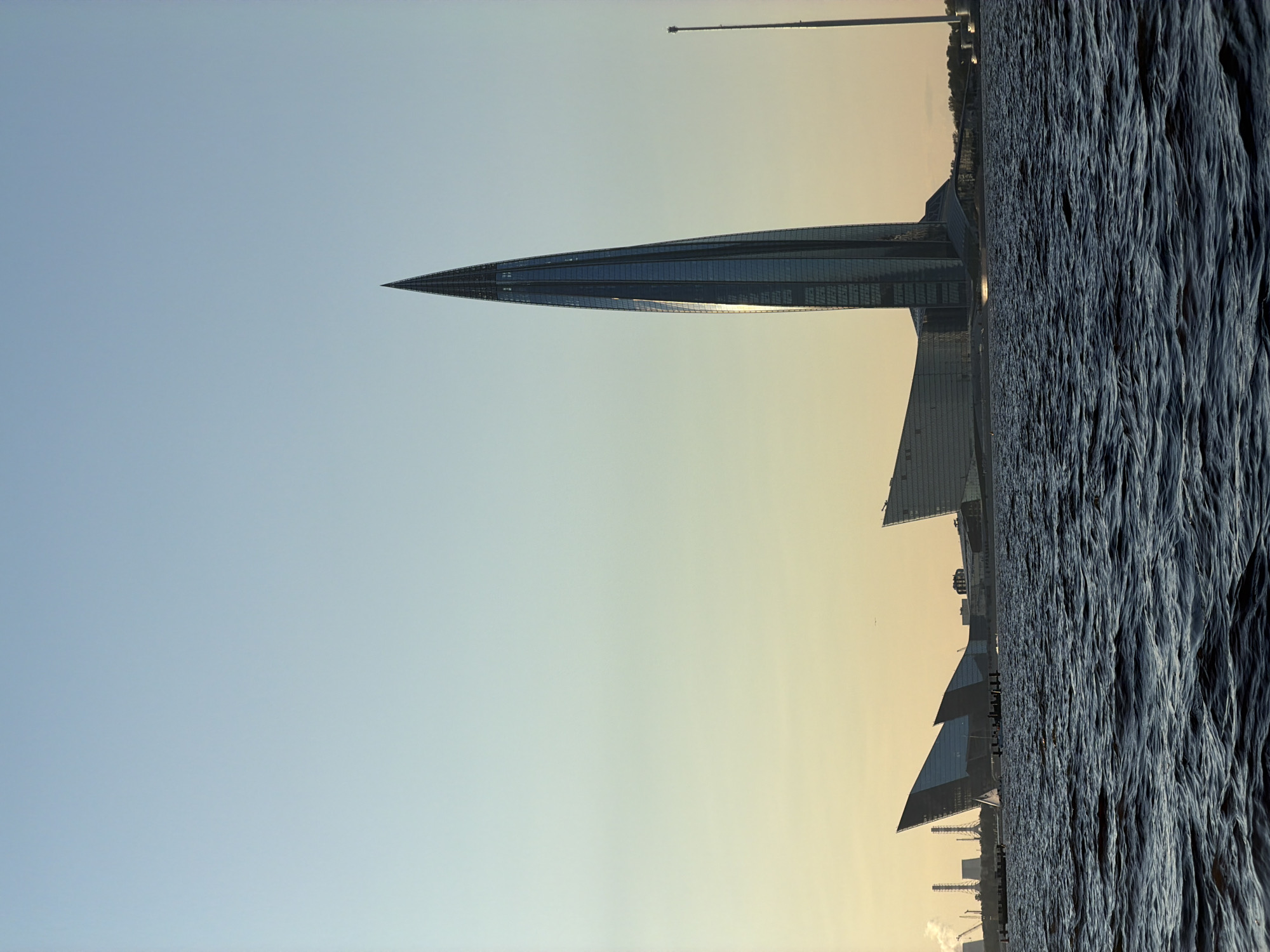 The image shows the Lakhta Center in Saint Petersburg, Russia, a striking skyscraper characterized by its pointed, twisting form. The building, with its glass facade reflecting the sky, dominates the right side of the frame. In the background, to the left of the Lakhta Center, are several other modern architectural structures with angular designs and some industrial chimneys emitting steam. The foreground is occupied by a body of dark, choppy water, likely the Gulf of Finland, with small white waves indicating movement. A thin, tall pole is visible to the right of the Lakhta Center, and a small boat with a white sail is on the water in the middle ground. The sky is a gradient of pale blue at the top, transitioning to a soft yellow hue near the horizon, suggesting either early morning or late evening light. No people are clearly visible in the scene.