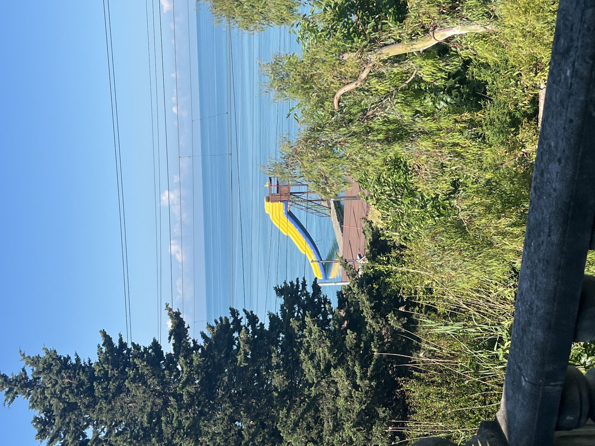 A bright blue sky with a few scattered white clouds hovers over a calm, turquoise sea. Several power lines stretch horizontally across the sky. In the mid-ground, a vibrant yellow and blue water slide stands on a wooden platform, its entrance near the top and its exit pointing towards the sea. The slide is surrounded by lush green trees and vegetation. In the foreground, a stone railing runs horizontally across the bottom of the image. The overall scene suggests a sunny day at a coastal location.