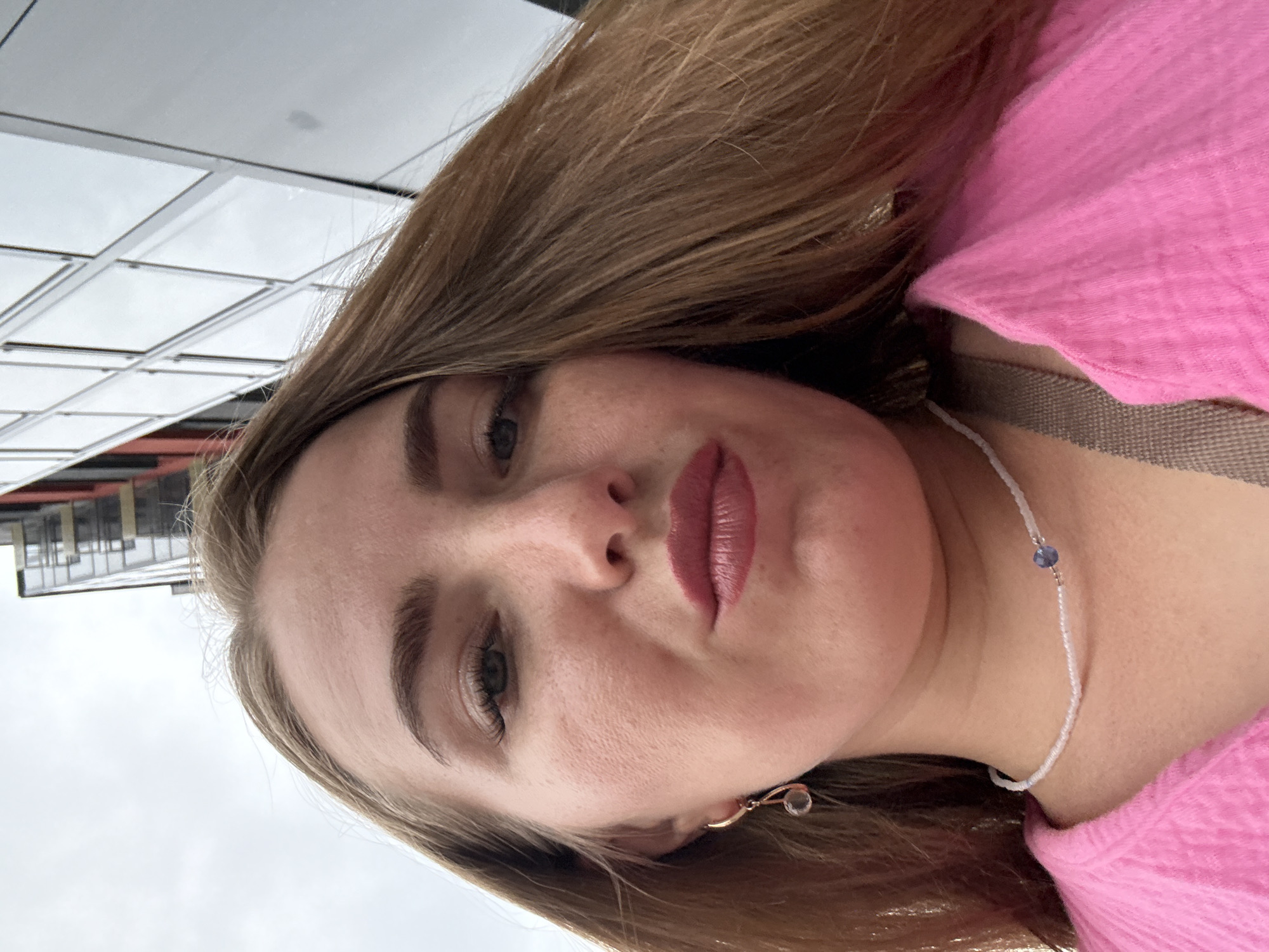 A low-angle, close-up, point-of-view selfie of a young woman with long, blonde hair, wearing a pink top and a beaded necklace. She is looking directly at the camera with parted lips. Her right eye is clearly visible with mascara and eyeshadow. A light-colored earring with a teardrop-shaped pendant hangs from her earlobe. The background consists of a cloudy sky and a modern building with glass windows, characteristic of an urban environment. The building's reflection is visible in the windows. The scene is outdoors.