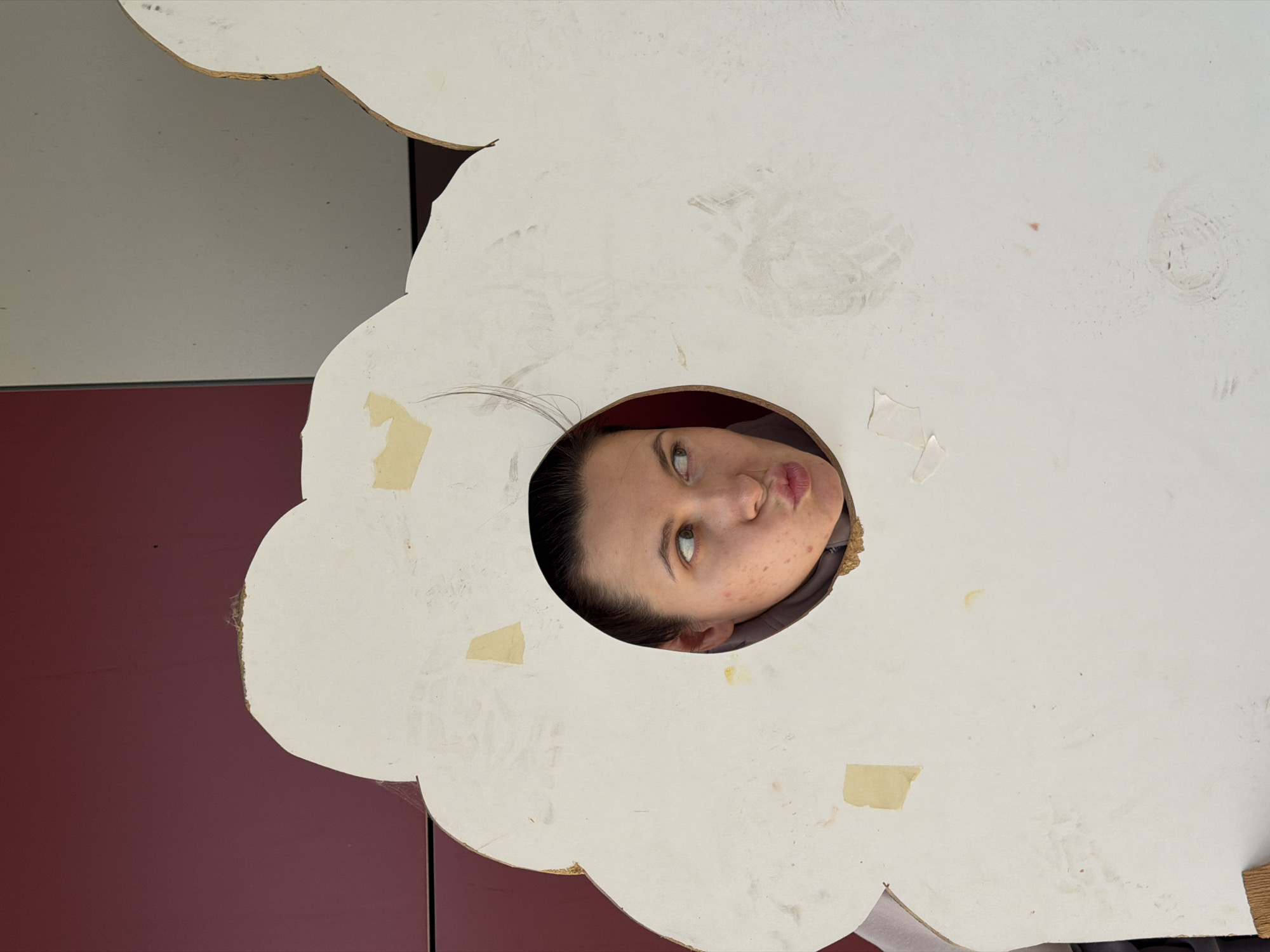 A person is peeking through a cutout in a white, cloud-shaped object. The person's face is tilted upwards and to the left, with their lips pursed. They have light skin with some blemishes and are looking off to the side with their eyes wide open. A few strands of dark hair are visible near their forehead. The cloud-shaped object is made of a white material that appears to be cardboard or foam board, with some light staining and scuff marks on its surface. There are also pieces of tape attached to the object. The background consists of a dark red surface on the left and a lighter, gray surface on the top. The scene suggests a playful or curious pose, possibly at an outdoor event or photo opportunity. The lighting is bright, indicating it is daytime. The location context is Красный Октябрь, Russia. No text is visible in the image.