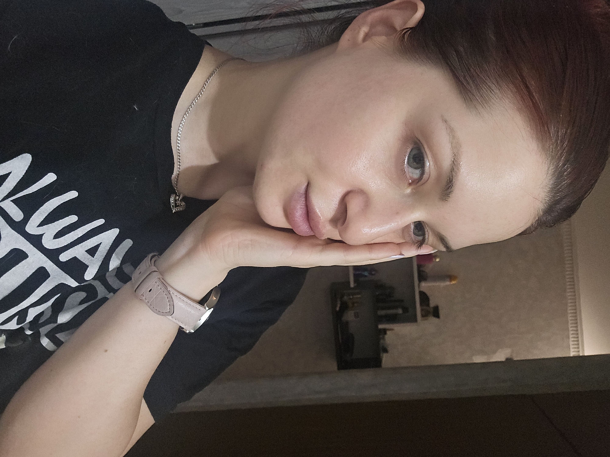 A woman is resting her face on her hand, looking slightly down and to the right of the frame. She has fair skin, light eyes, and brown hair pulled back from her face. She is wearing a black t-shirt with white lettering, a silver necklace with a heart pendant, and a light pink watch. The background appears to be an interior setting, with a shelf visible behind her. The photo is taken from an elevated angle and the image is rotated slightly counter-clockwise. There are no discernible details to identify a specific location in Burgas, Bulgaria.