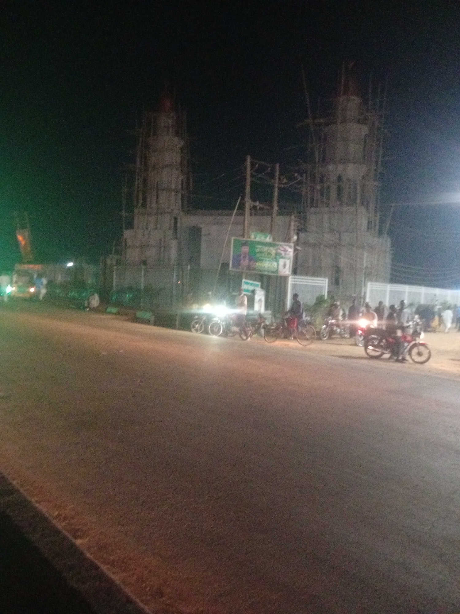 The image depicts a nighttime scene in Tundun Wada West, Nigeria. A large, unfinished structure, likely a mosque given its minaret-like towers and scaffolding, dominates the background. The structure is illuminated by artificial lights, casting a dim glow on the surrounding area.
In the foreground and mid-ground, a street is visible with several motorcycles and bicycles parked or in motion. A few people are gathered around the vehicles, suggesting a social or commercial hub. Some vehicles appear to be trucks or vans. The street itself is paved and dimly lit, with the headlights of vehicles creating streaks of light.
The sky is dark, indicating it is nighttime. The weather appears clear, with no visible signs of rain or fog. The overall atmosphere is somewhat chaotic due to the construction and the presence of numerous vehicles and people.
One notable detail is a large billboard on the left side of the unfinished structure, displaying an image of a man and some text in what appears to be Arabic or a similar script. The text is too blurry to read. Another billboard or sign is visible further to the right, also with unreadable text.
The image seems to capture a moment in the everyday life of Tundun Wada West, highlighting ongoing development and local activity at night.