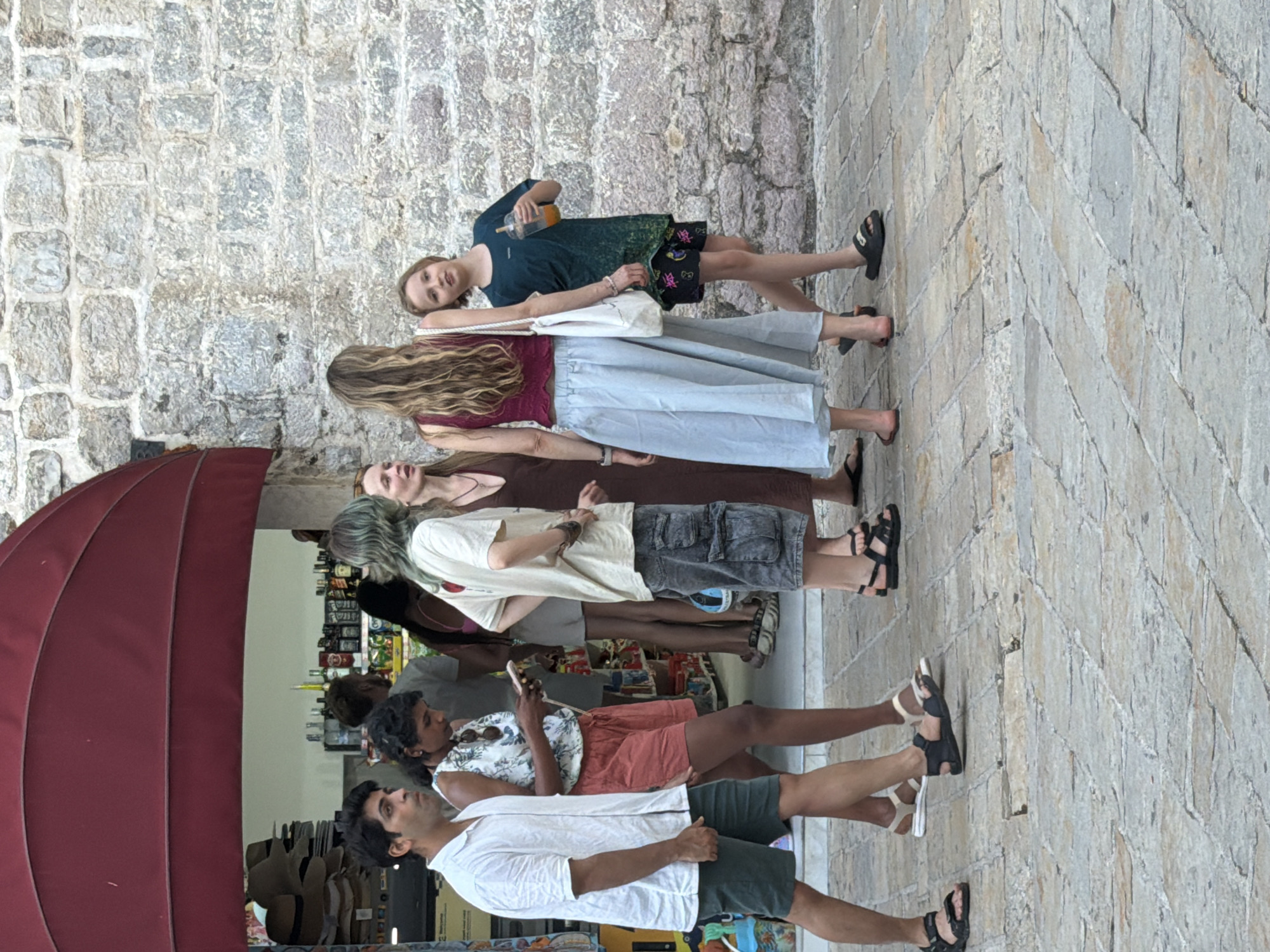 A group of six people, consisting of five adults and one child, are walking and standing outside a shop in Budva, Montenegro. The shop has a red awning and displays hats and bottles in its window. The ground is paved with cobblestones. A man in a white shirt and green shorts walks on the left. Next to him, a woman in a floral top and coral shorts looks at her phone. Behind them, a woman with green hair and a woman in a brown dress stand talking. To the right, a woman with long blonde hair in a burgundy top and a light blue skirt stands with a white bag. A child in a dark green shirt and patterned shorts, holding an orange drink, stands to the far right.