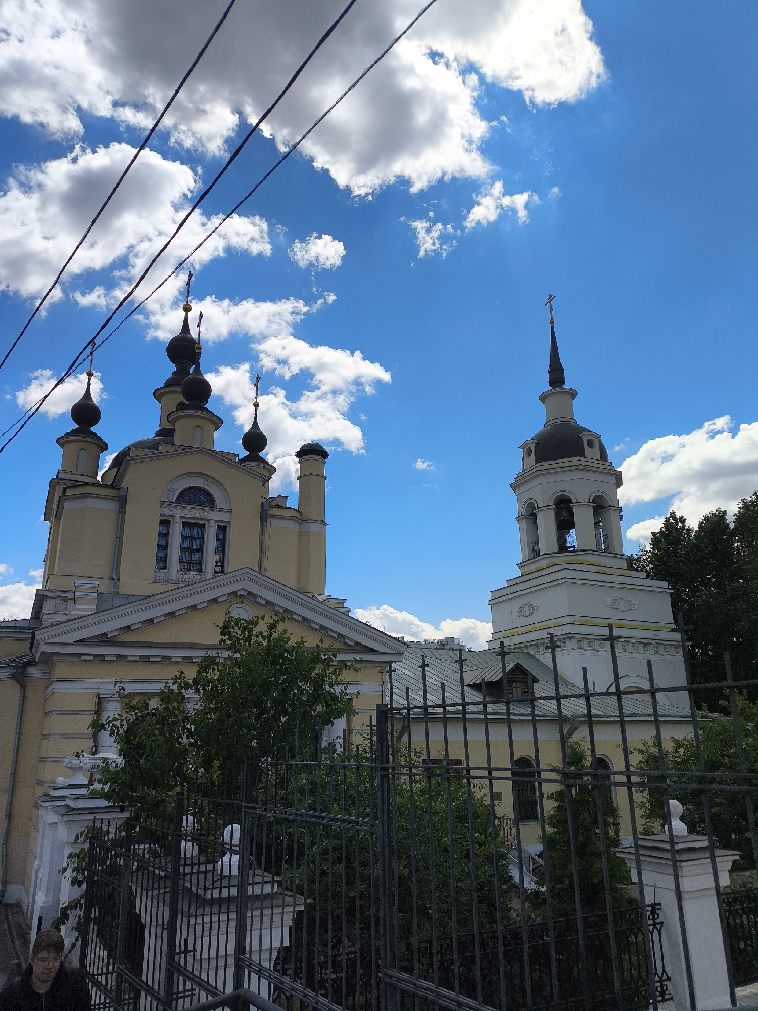 The image shows a yellow church with multiple domes and a white bell tower, set against a bright blue sky with fluffy white clouds. Overhead, several dark electrical wires stretch across the frame, obscuring parts of the sky. In the foreground, a black metal fence partially blocks the view of the church complex and surrounding greenery.
The church itself features a light yellow facade with ornate architectural details. Several dark, bulbous domes topped with crosses rise from the main structure, while the bell tower is predominantly white with a dark, conical roof and visible bell openings. Lush green trees and bushes are visible behind the fence and around the buildings.
The lighting suggests it is daytime, likely with bright sunshine filtering through the clouds. The overall scene conveys a sense of religious architecture and a somewhat urban or suburban setting, given the presence of the electrical wires and fencing. There is no visible text in the image.