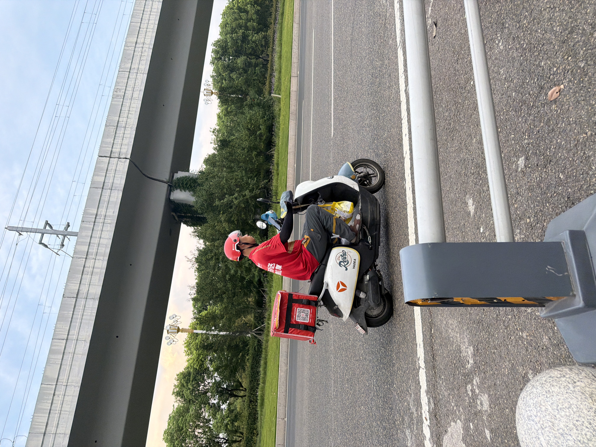 A delivery person on a scooter is captured in profile, riding on a road. The delivery person is wearing a red shirt, a red helmet with a clear visor, and grey pants. A large red insulated delivery box is mounted on the back of the scooter. The scooter is white with black accents and has a logo that reads "The Retro YADEA". The scooter is positioned on the right lane of a multi-lane road, moving towards the right of the frame. In the background, there is a large elevated concrete structure, likely a bridge or overpass, with power lines running above it. Lush green trees and some streetlights are visible on the side of the road. The sky is overcast with a hint of sunset colors. In the foreground, a grey bollard with yellow accents and a concrete sphere are visible on the side of the road. The image was taken in Jiangjin, China, as indicated by the delivery box's labeling which includes Chinese characters, common in this region for food delivery services.