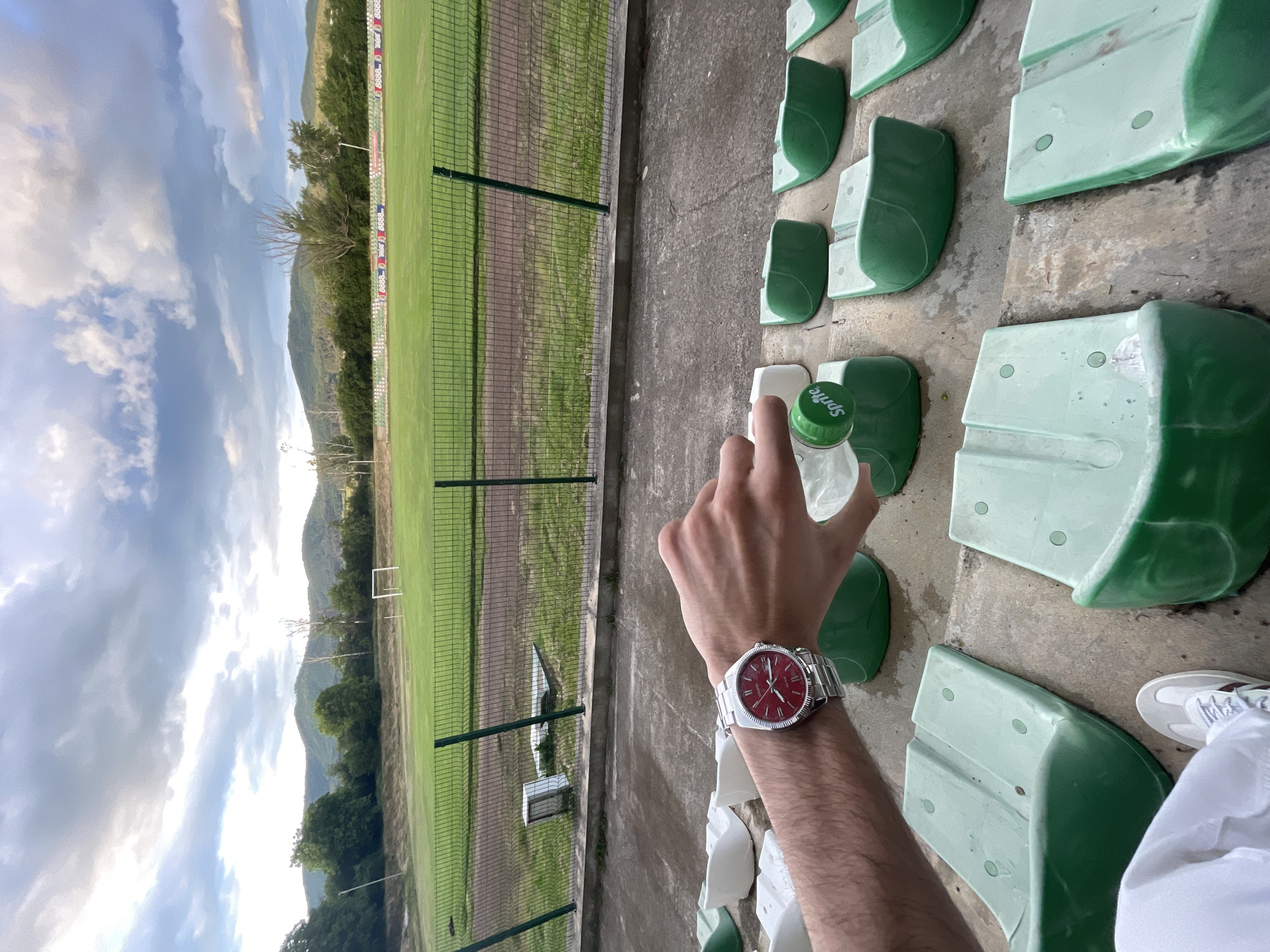 The image captures a first-person perspective from the stands of a sports stadium in Ihtiman, Bulgaria, looking out over a football field towards a mountainous backdrop.
In the immediate foreground, a person's right arm and hand are visible. The hand holds a plastic bottle with a green cap, clearly branded "Sprite." A silver-toned wristwatch with a prominent red dial and a fluted bezel is worn on the wrist. To the lower right, part of a white sneaker and white clothing (likely pants or shorts) are visible.
The seating area consists of rows of light green plastic seats set on a concrete surface. Beyond the seating, a metal fence separates the stands from a well-maintained green grass football pitch. Two white goalposts can be seen on the field. Along the perimeter of the field, near the fence, a red advertisement banner with repeated "8888" logos is discernible.
The background features a range of green, tree-covered mountains under a partly cloudy sky. The clouds are a mix of white and darker grey, suggesting either an overcast day or the approaching evening. The overall lighting is soft and natural. The scene appears calm, possibly capturing a moment during a game, practice, or simply observing the field.