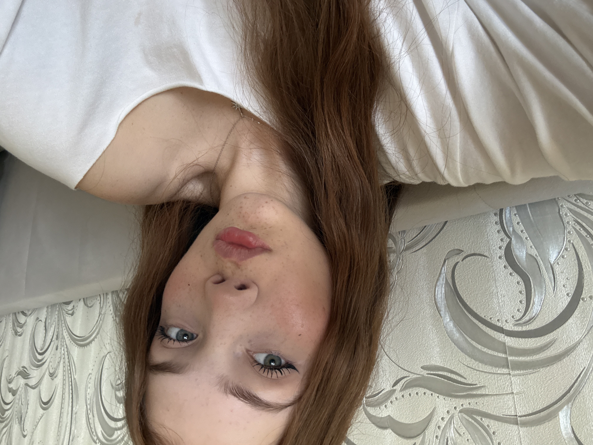 This media file presents a close-up portrait of a young woman, photographed from an overhead angle and rotated 90 degrees counter-clockwise, causing her to appear upside down. She has reddish-brown hair, fair skin adorned with visible freckles across her nose and cheeks, and striking blue/grey eyes. Her lips are naturally pink and slightly parted. She appears to be wearing light eye makeup, including mascara. Around her neck, a delicate silver chain necklace is visible. She is dressed in a simple white T-shirt or top.
The background consists of a patterned wall, likely wallpaper, featuring ornate silver and grey swirling, floral-like designs against a lighter, textured surface. A section of white bedding or a pillow is also visible on the right side of the frame, near her hair and shoulder. The lighting is soft and diffused, indicating an indoor setting. Her expression is neutral and calm, with a direct gaze towards the camera. There is no specific activity or event taking place, other than the casual portraiture. No visible text is present in the image. The scene conveys a relaxed, intimate atmosphere.