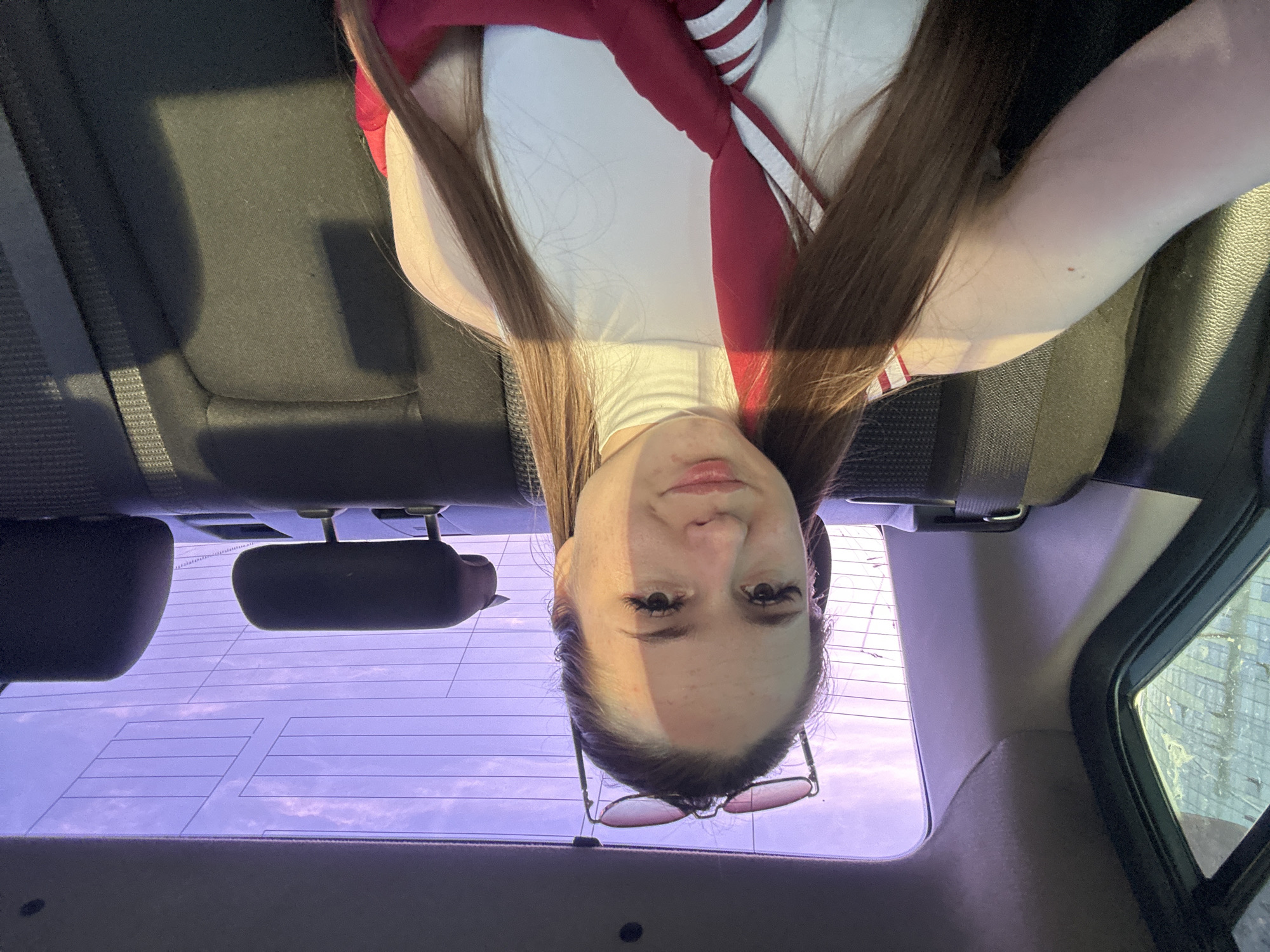 A young adult female with fair skin and long, straight, brown hair is positioned in the rear passenger seat of a vehicle, taking a selfie. She wears a white, sleeveless, high-neck top and a red and white striped garment draped across her chest and left shoulder. Sunglasses are resting on top of her head. Sunlight casts a distinct shadow diagonally across her face and body from the upper left. The vehicle's interior features dark fabric seats and gray-brown plastic trim. Through the rear window, a purplish-pink sky with faint clouds is visible, along with horizontal defroster lines and what appear to be antenna patterns on the glass. A partial view of an exterior building with horizontal elements is visible through the left rear window. The location is Unknown, Unknown.